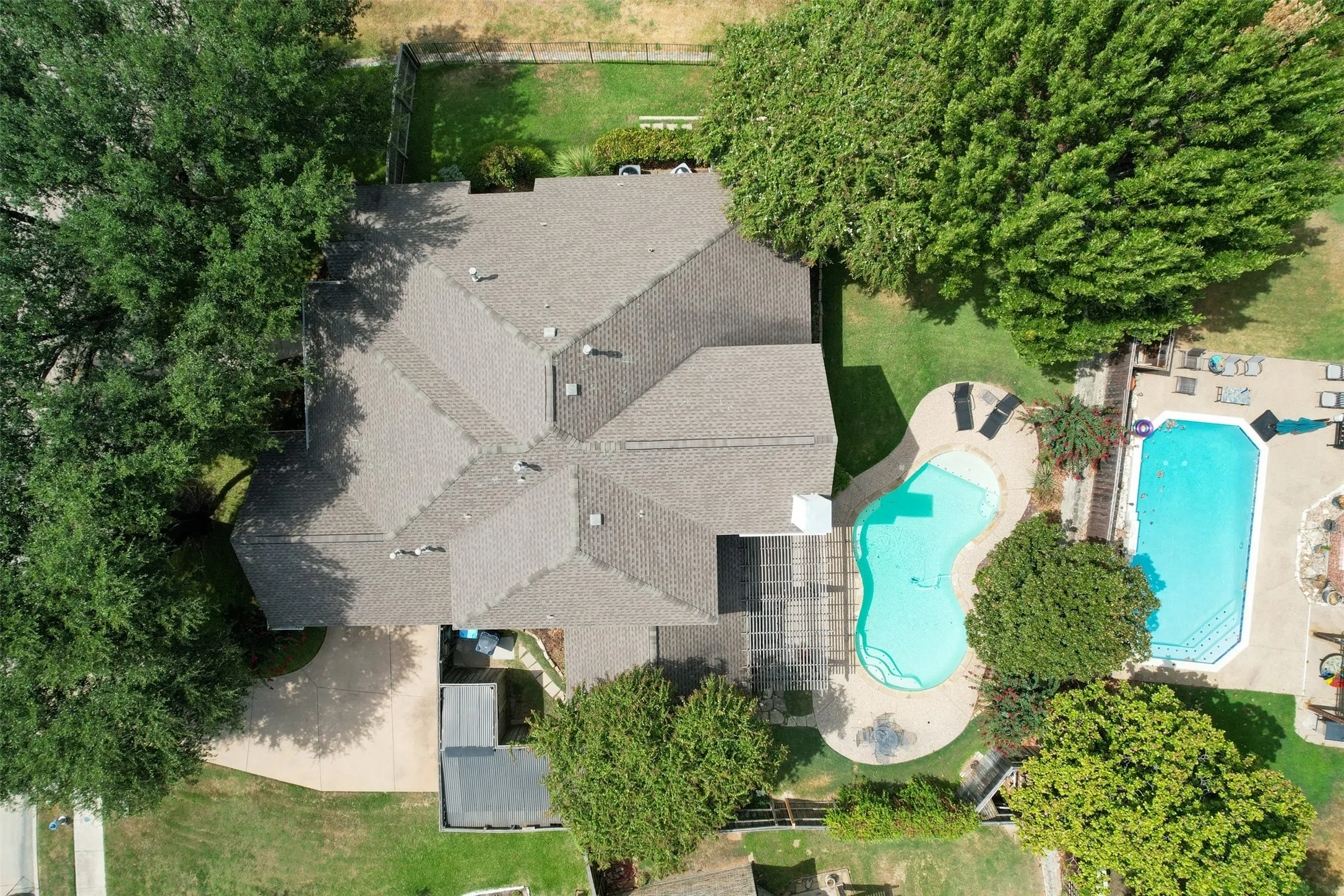 Two distinct swimming pools with surrounding hardscaping, a multi-pitched shingle roof, established trees, and a grassy area