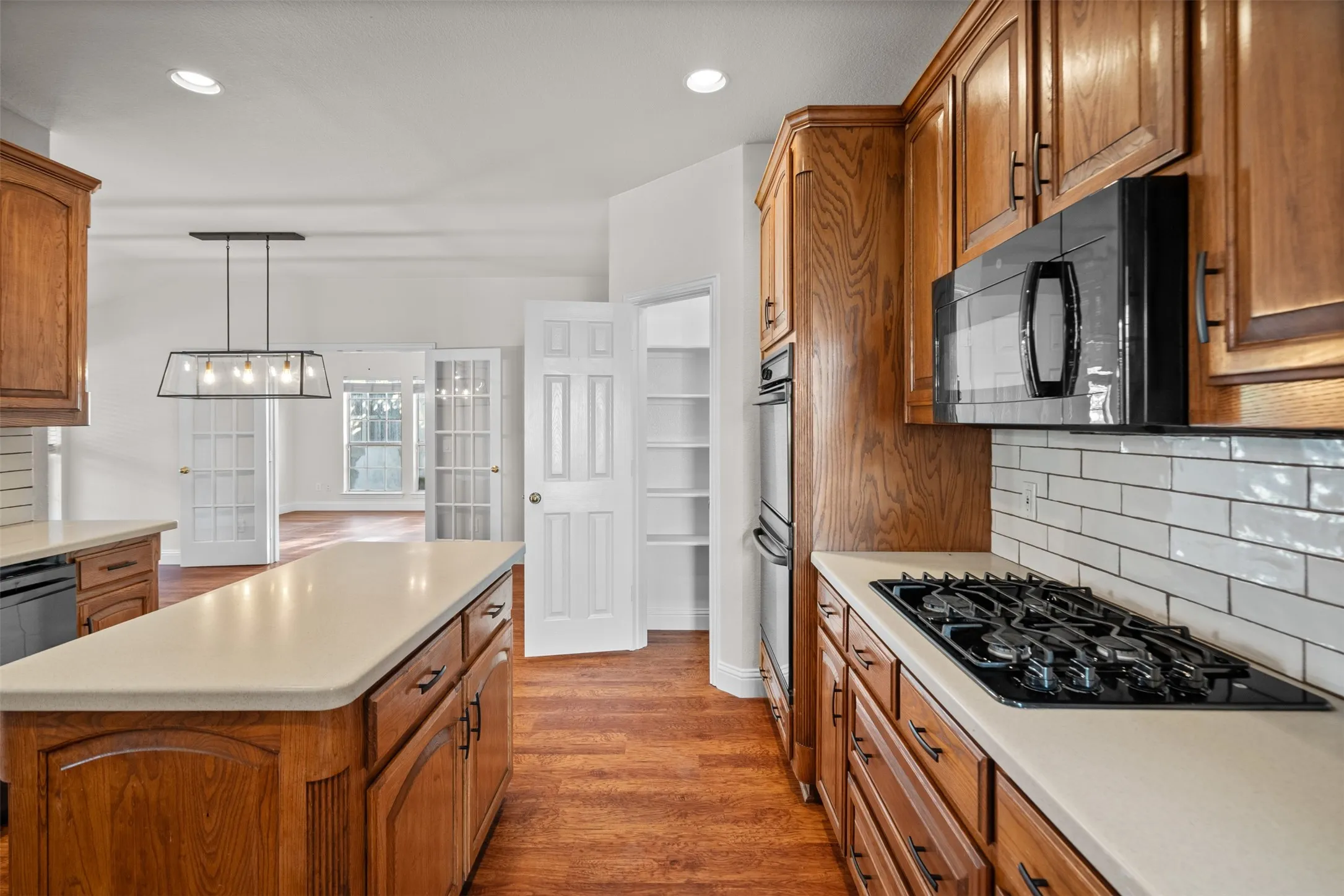 Wood-finish flooring, wood cabinetry with black hardware, light-toned countertops, white subway tile backsplash, and a built-in gas cooktop