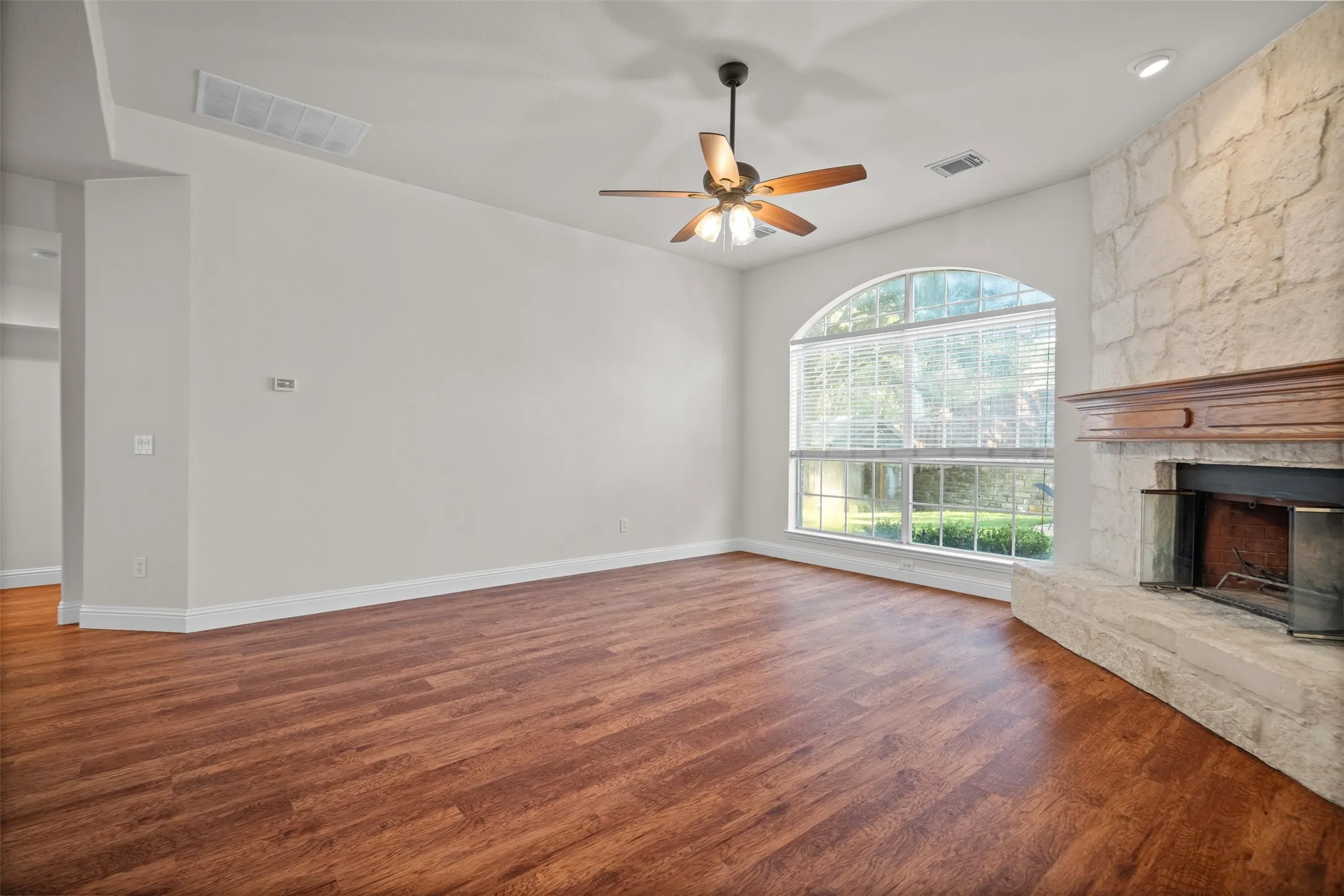Stone-clad fireplace with wood mantel, arched window, wood-finish flooring, and ceiling fan with light fixture