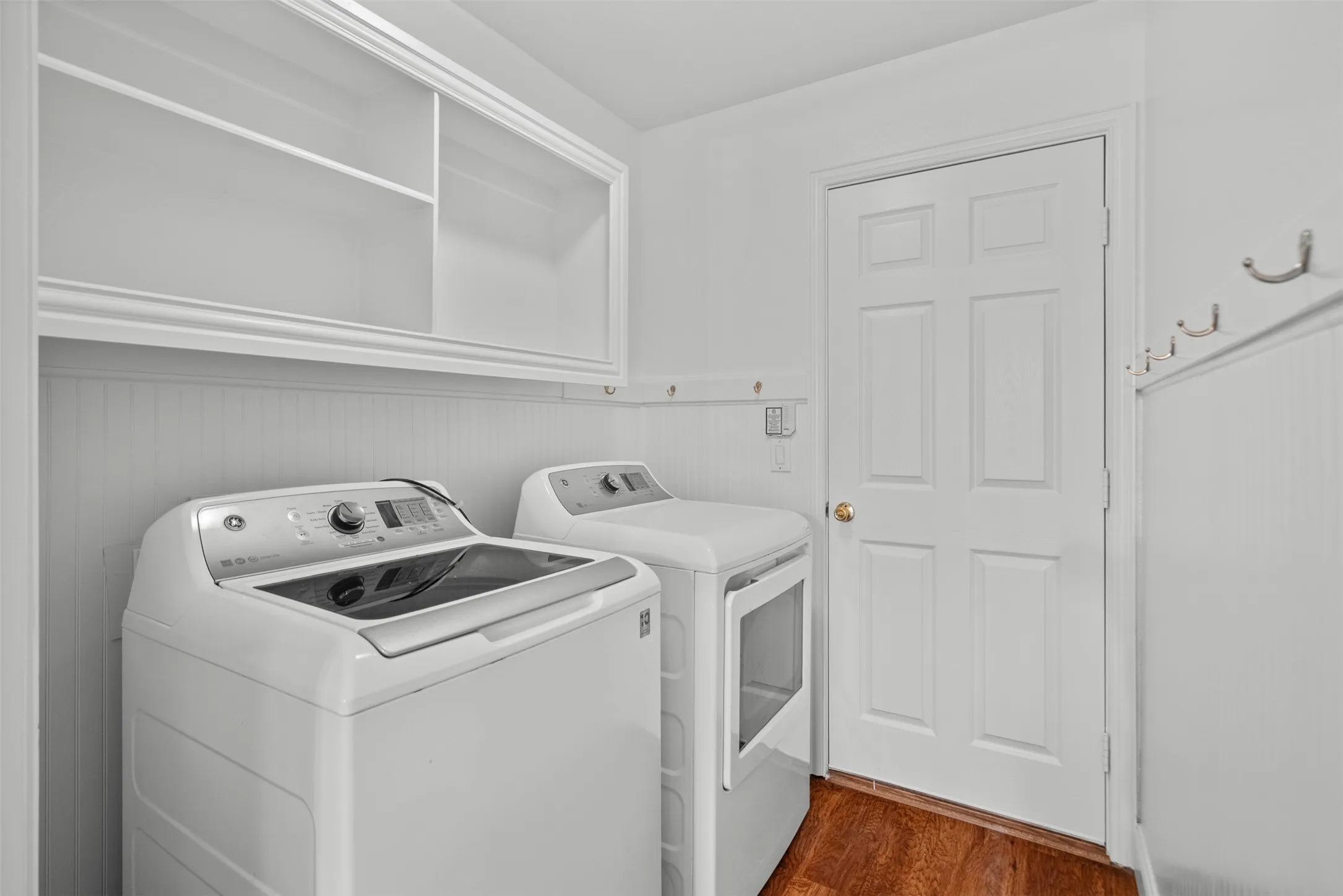 Laundry area with overhead white cabinetry, beadboard wainscoting, wall-mounted hooks, and wood-finish flooring