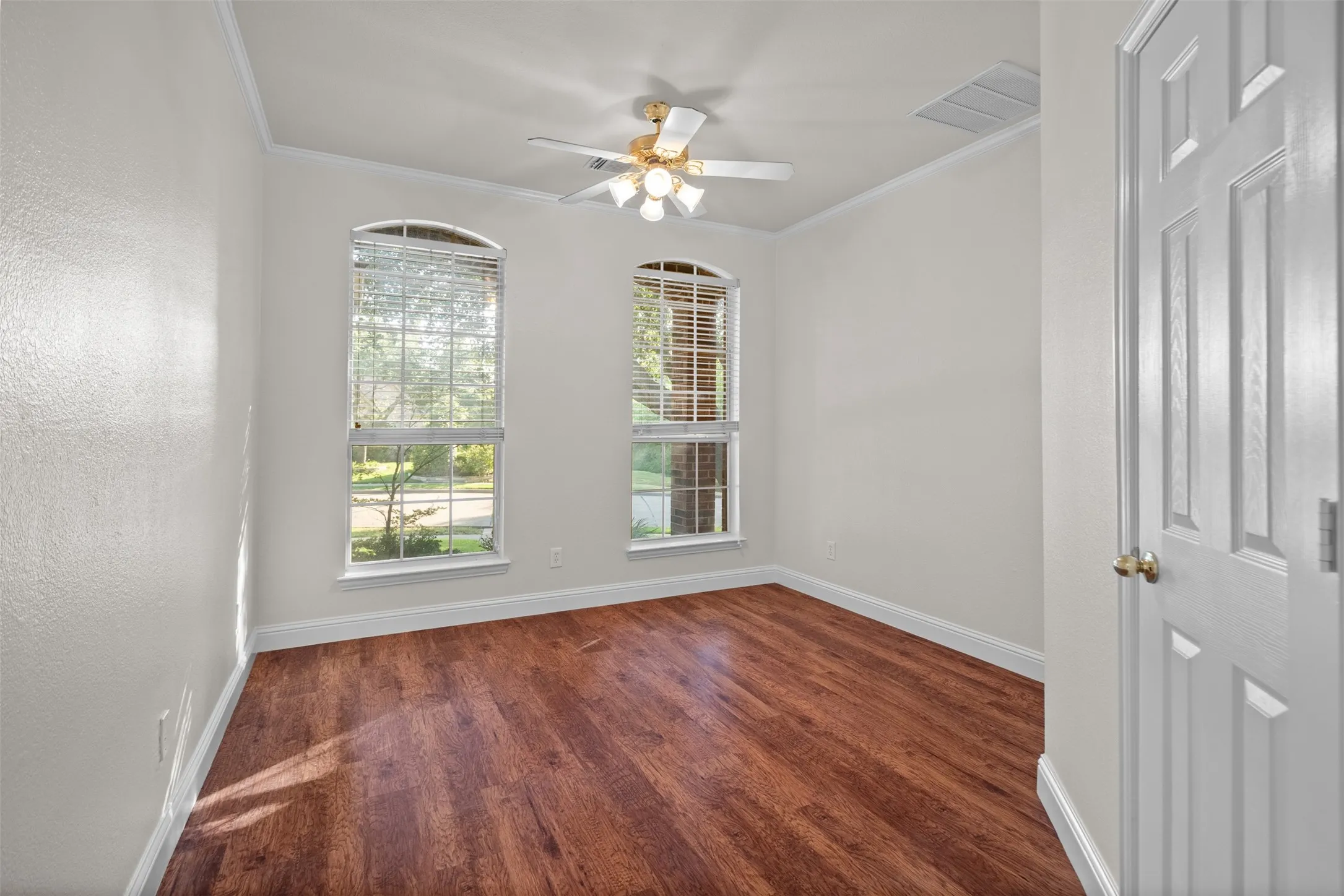 Wood-finish flooring, two arched windows with blinds, ceiling fan with light fixture, white paneled door, and crown molding