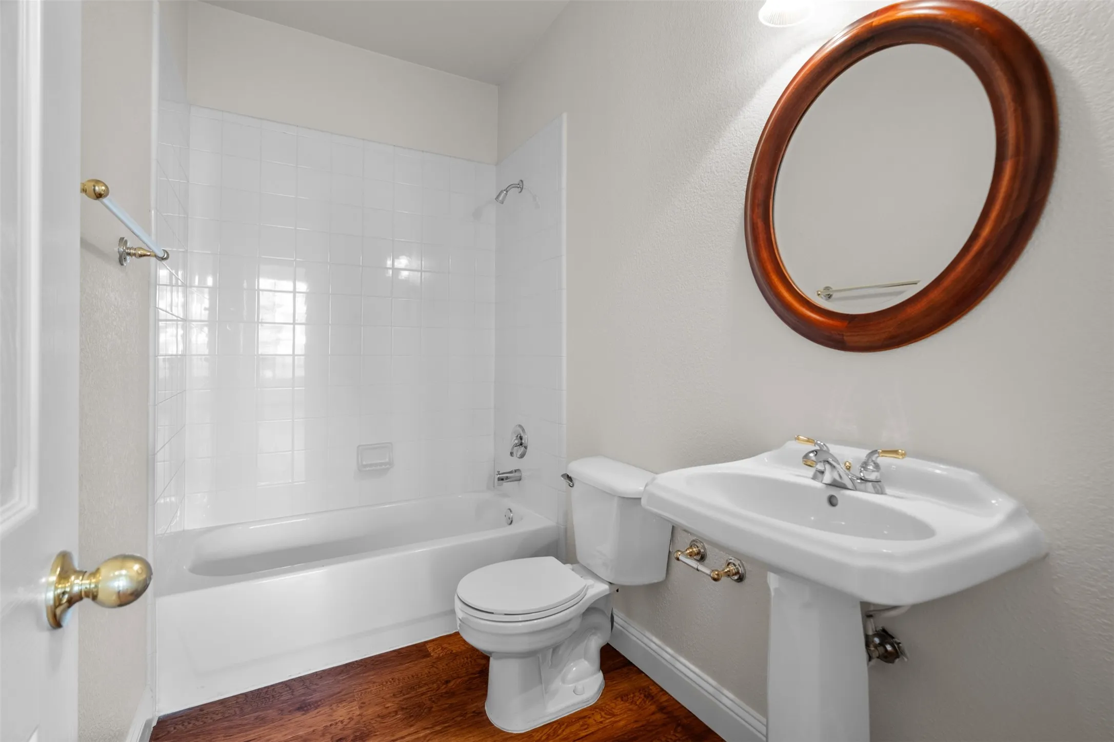 Pedestal sink with dual-handle faucet, oval wood-frame mirror, white bathtub with tile surround, and wood-finish flooring