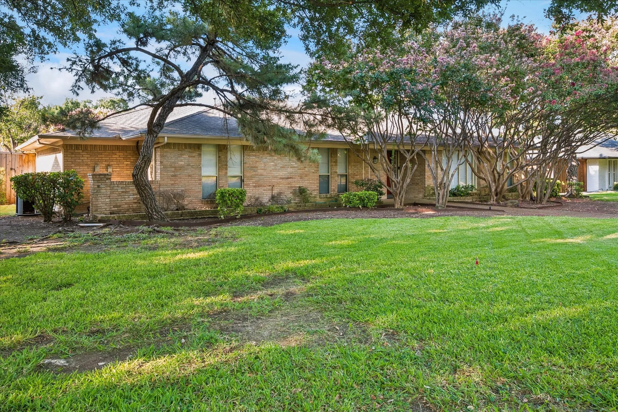 Single-story brick home with a manicured lawn and lush, tree-shaded landscaping.