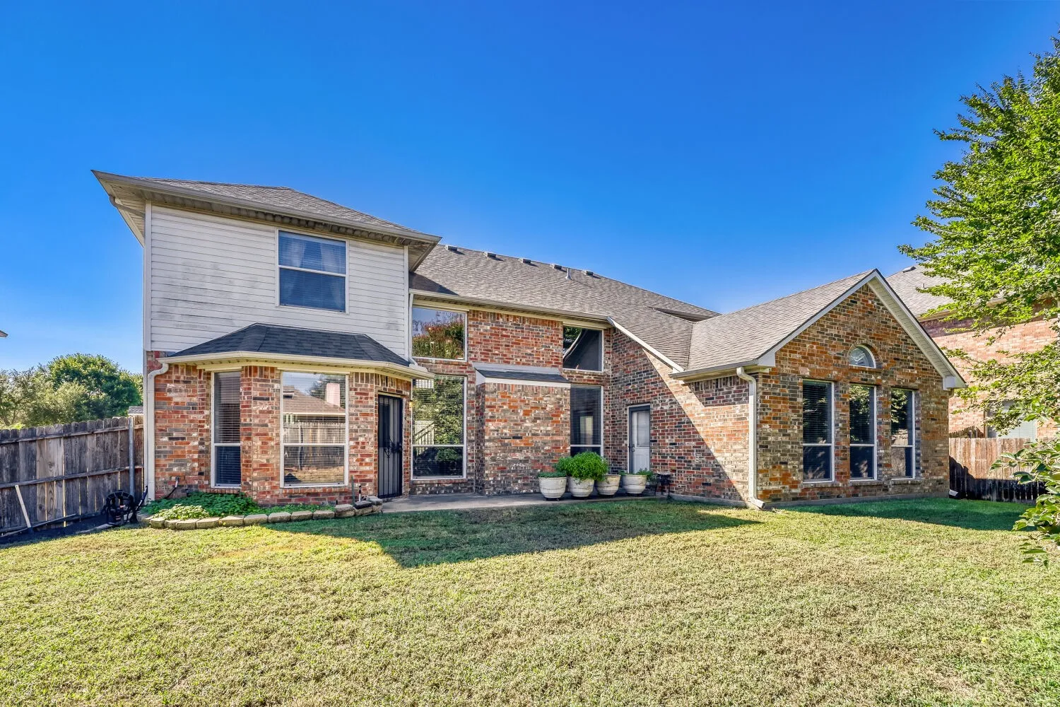 Expansive backyard with a manicured lawn, multi-level brick and siding exterior, multiple large windows, and a privacy fence