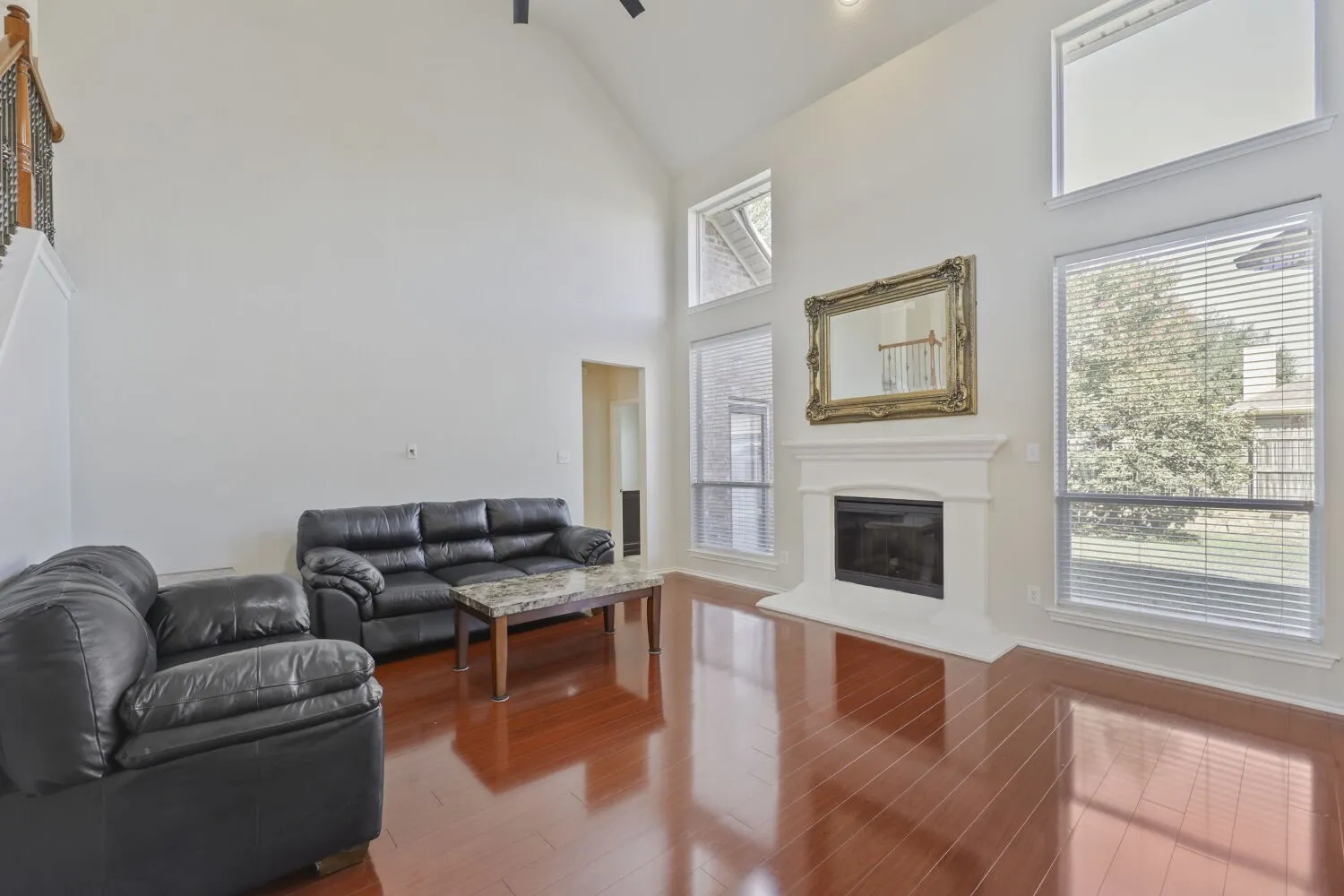 Vaulted ceiling with a ceiling fan, fireplace with a mantle, large windows with blinds, wood-finish flooring, and a staircase with an ornamental railing
