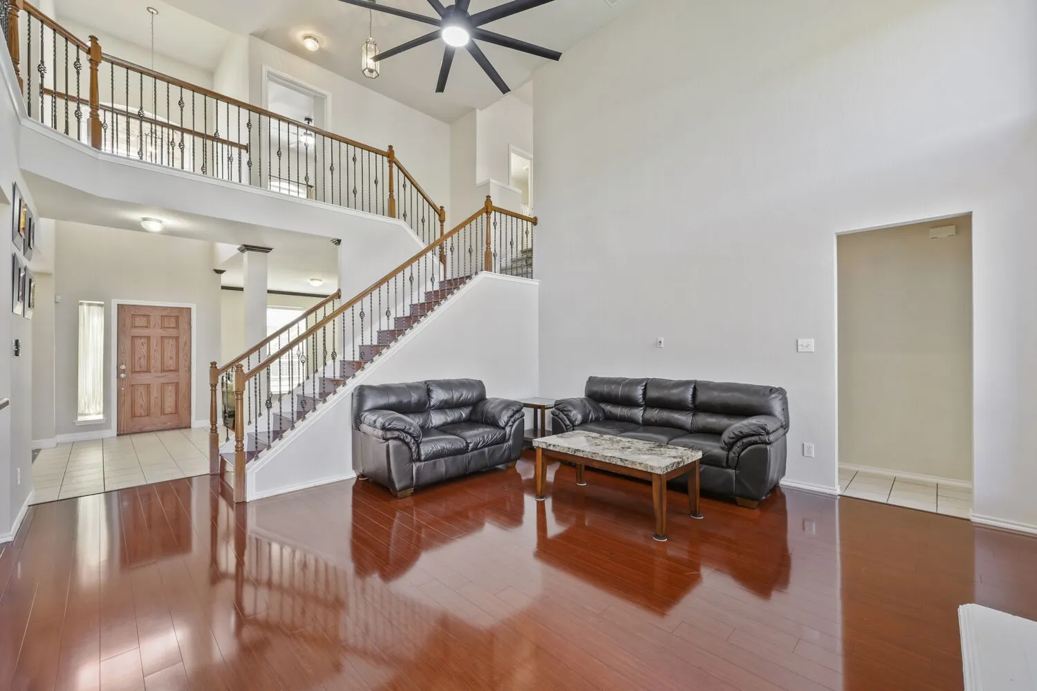 Two-story ceiling, wood-finish flooring, dual staircase with wrought iron balusters, and a ceiling fan with integrated lighting