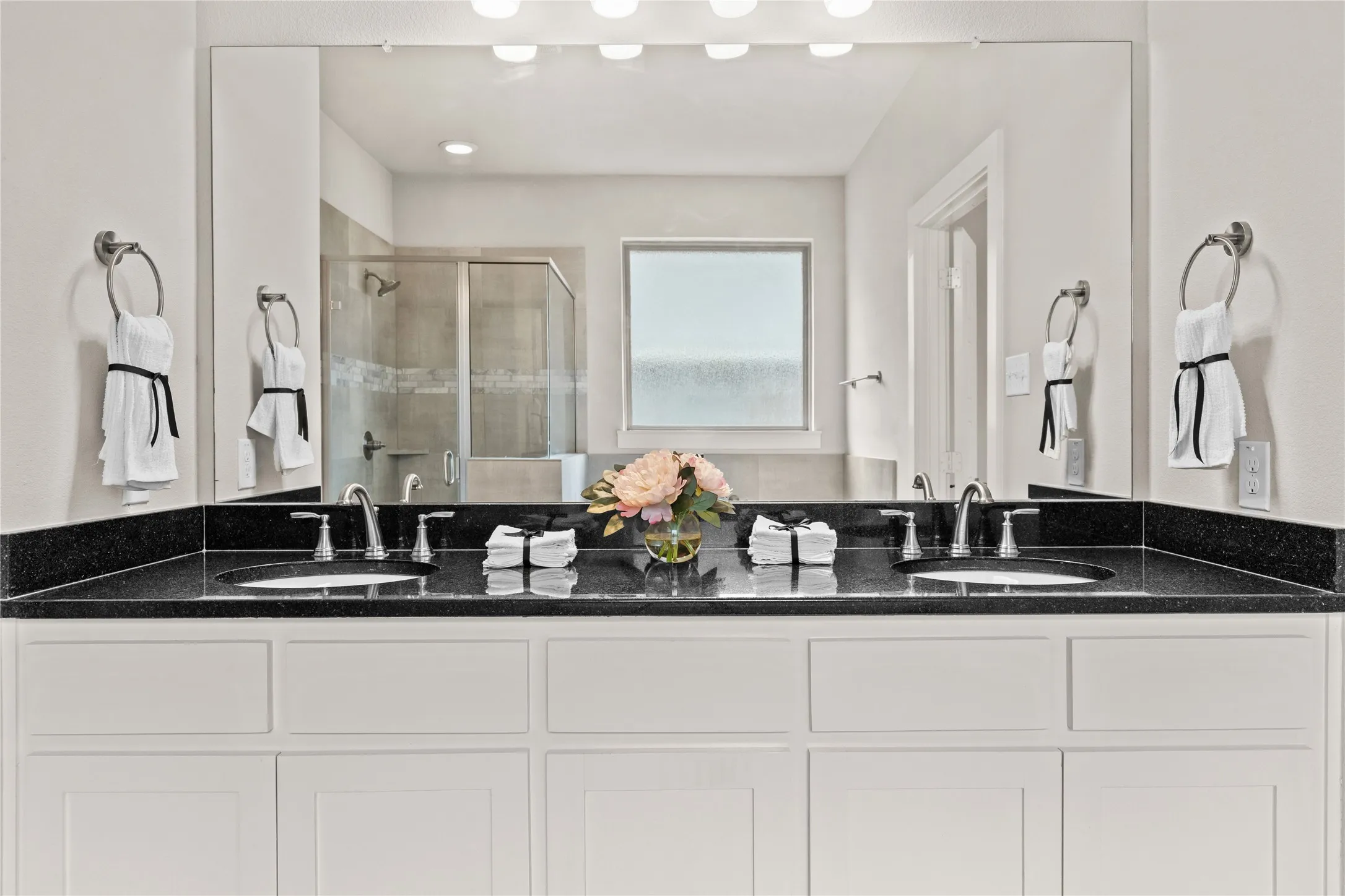 Dual vanity with black countertop, white shaker-style cabinetry, chrome fixtures, and an expansive mirror