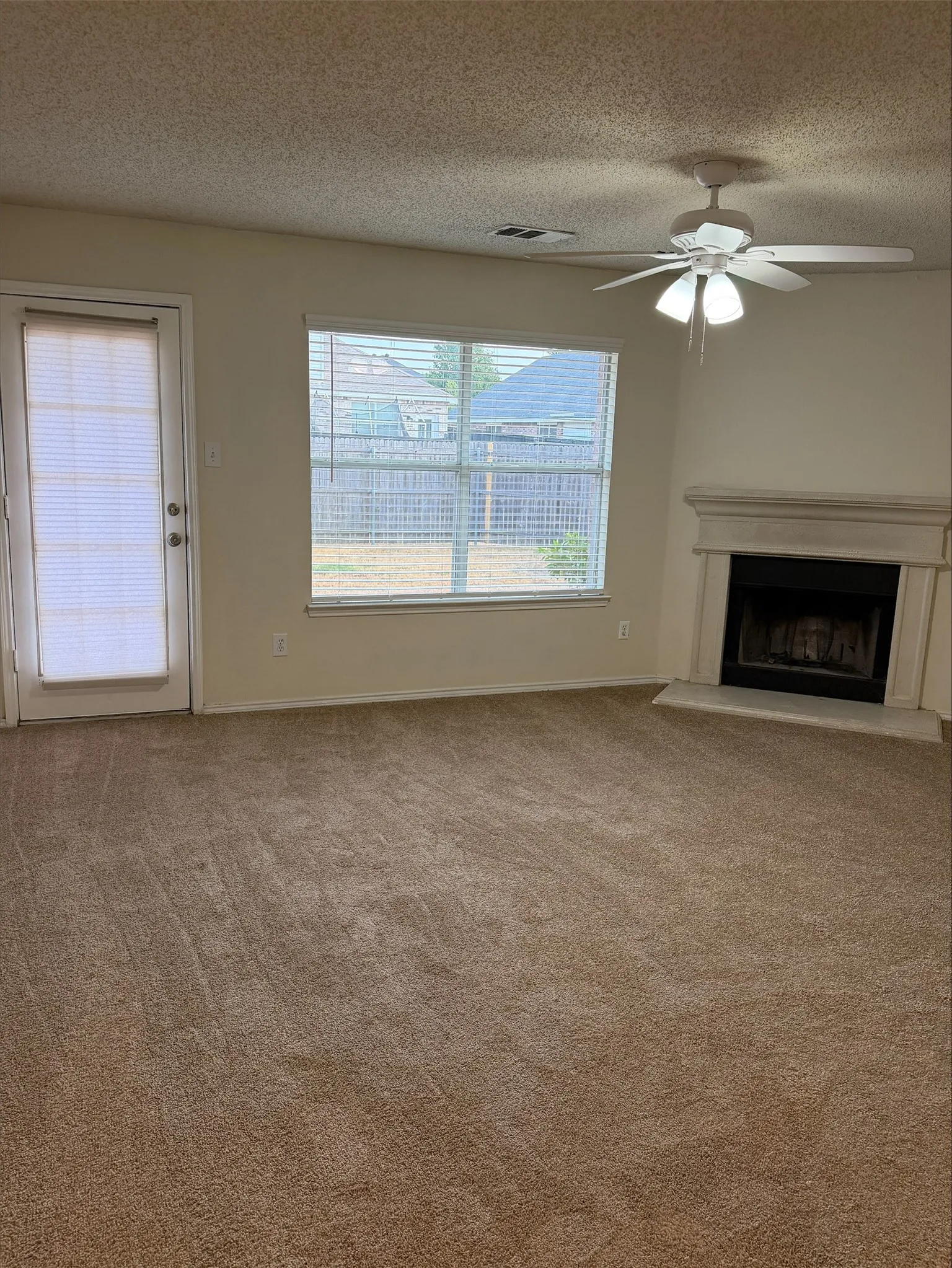 Neutral-toned carpeted flooring, corner-set fireplace with a light-colored mantel, window with horizontal blinds, and a ceiling fan with integrated lighting