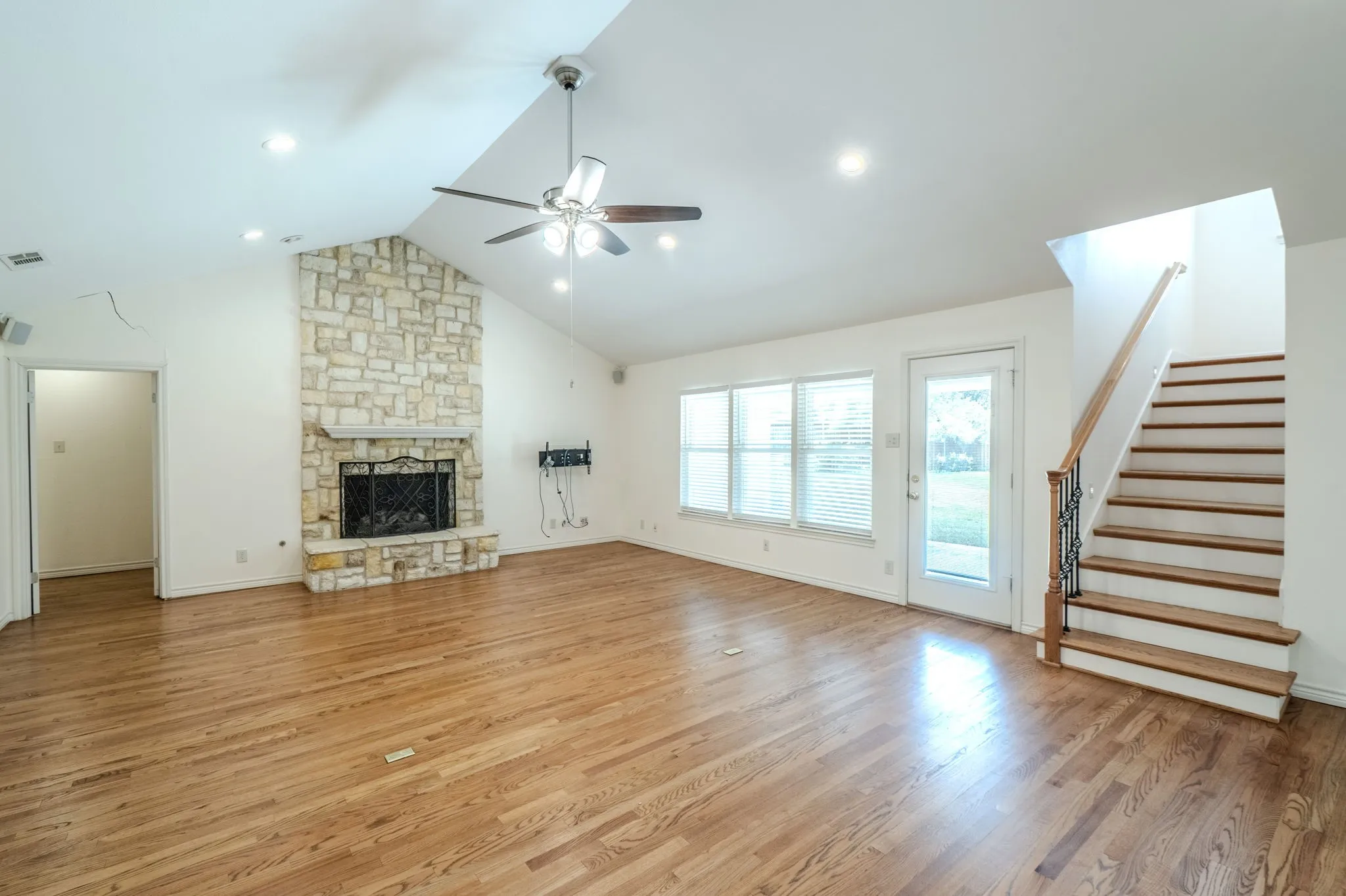 Vaulted ceiling, stone fireplace, hardwood flooring, large window banks, and a staircase with an ornamental iron railing