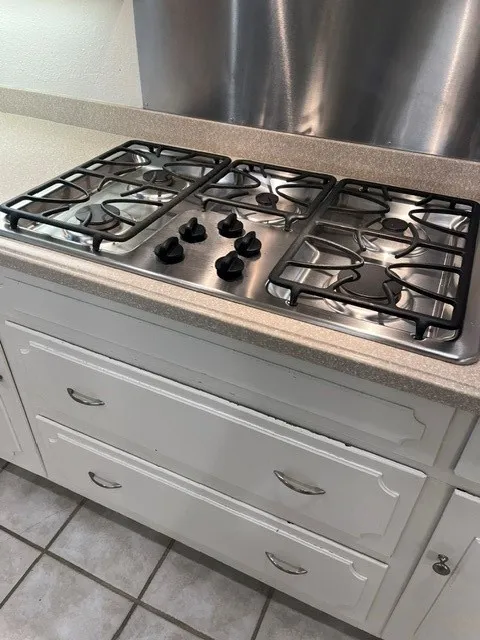 Built-in stainless steel cooktop with cast iron grates, light-toned countertop, stainless steel backsplash, and white cabinetry with drawer pulls