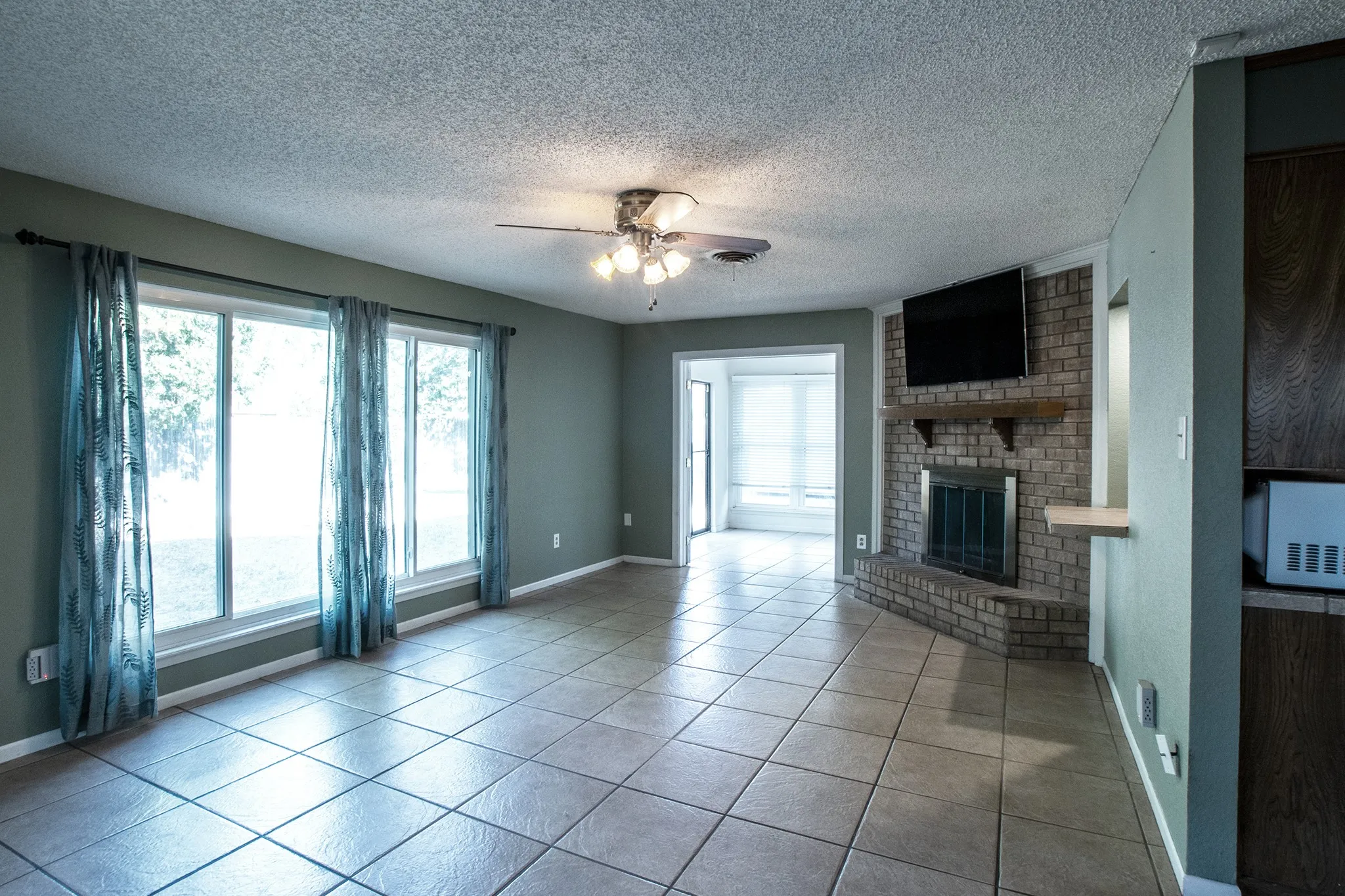 Tile flooring throughout, a brick-surround fireplace with a wood mantel, multiple windows, and a ceiling fan with integrated lighting