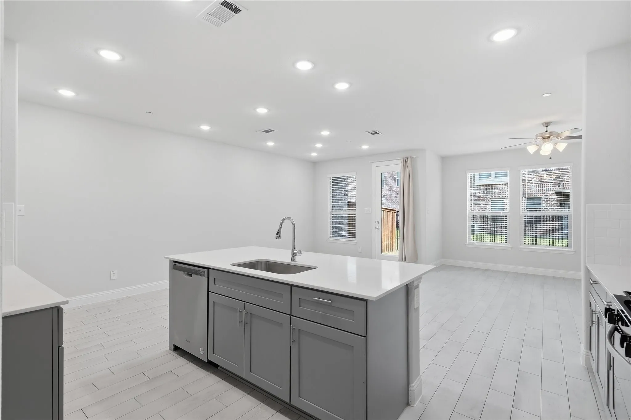 Kitchen island with an undermount sink and stainless steel faucet, stainless steel dishwasher, white quartz countertops, white tile backsplash, and light wood-finish flooring