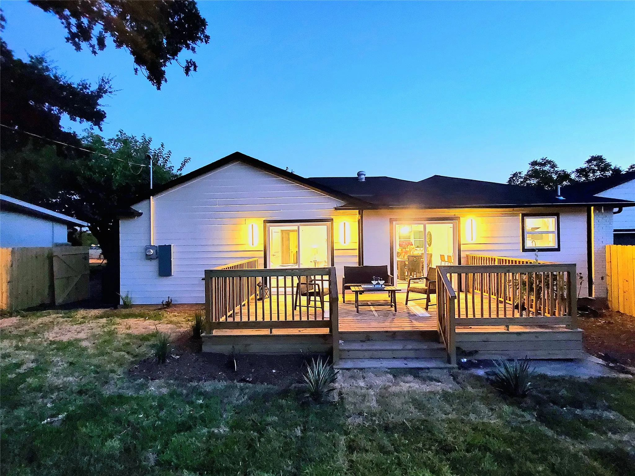 White exterior siding with black trim, wood deck, sliding glass doors, exterior lighting, and a fenced perimeter