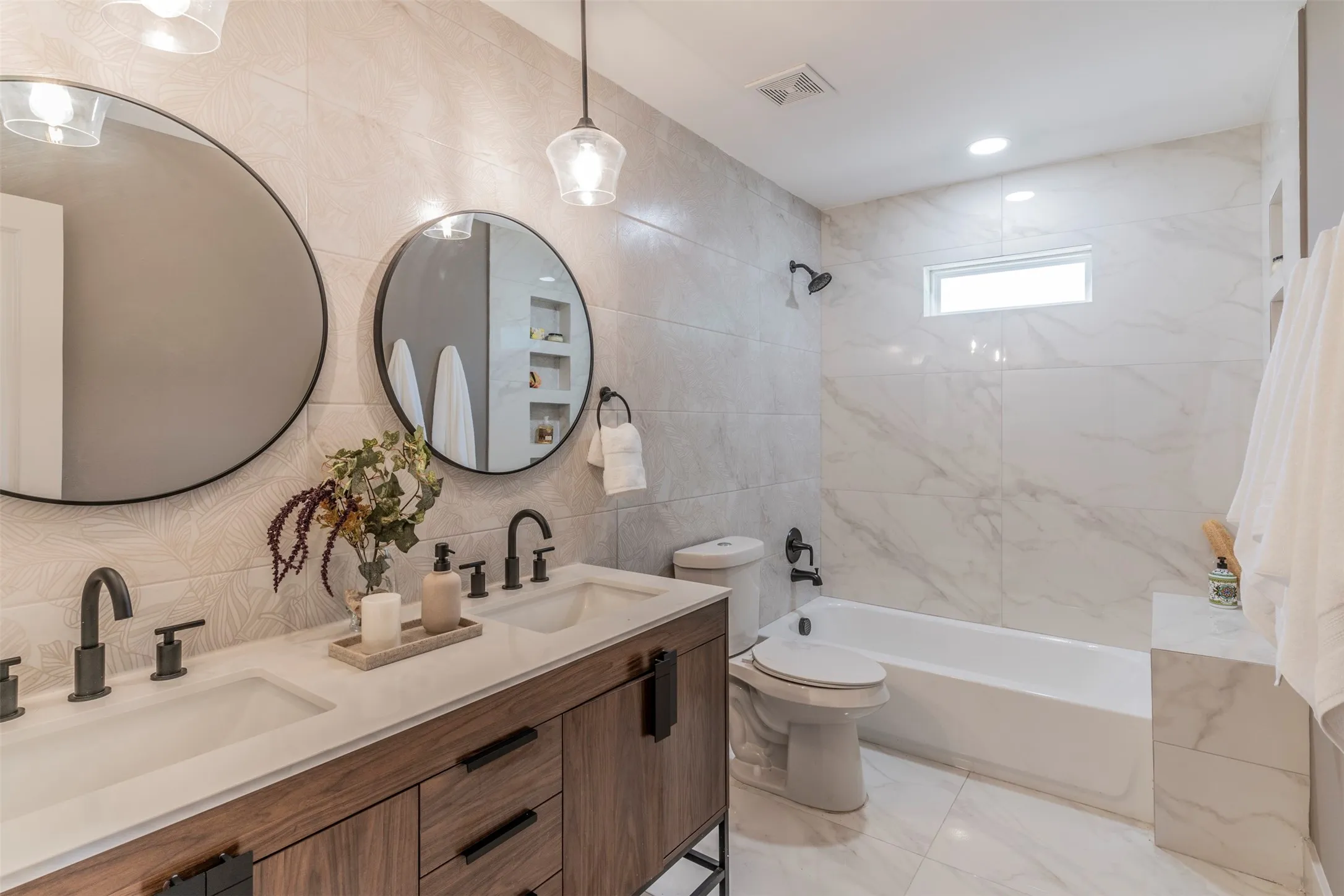 Dual round mirrors, a wood-finish double vanity with matte black hardware, a built-in tub, and marble-look tile flooring and walls