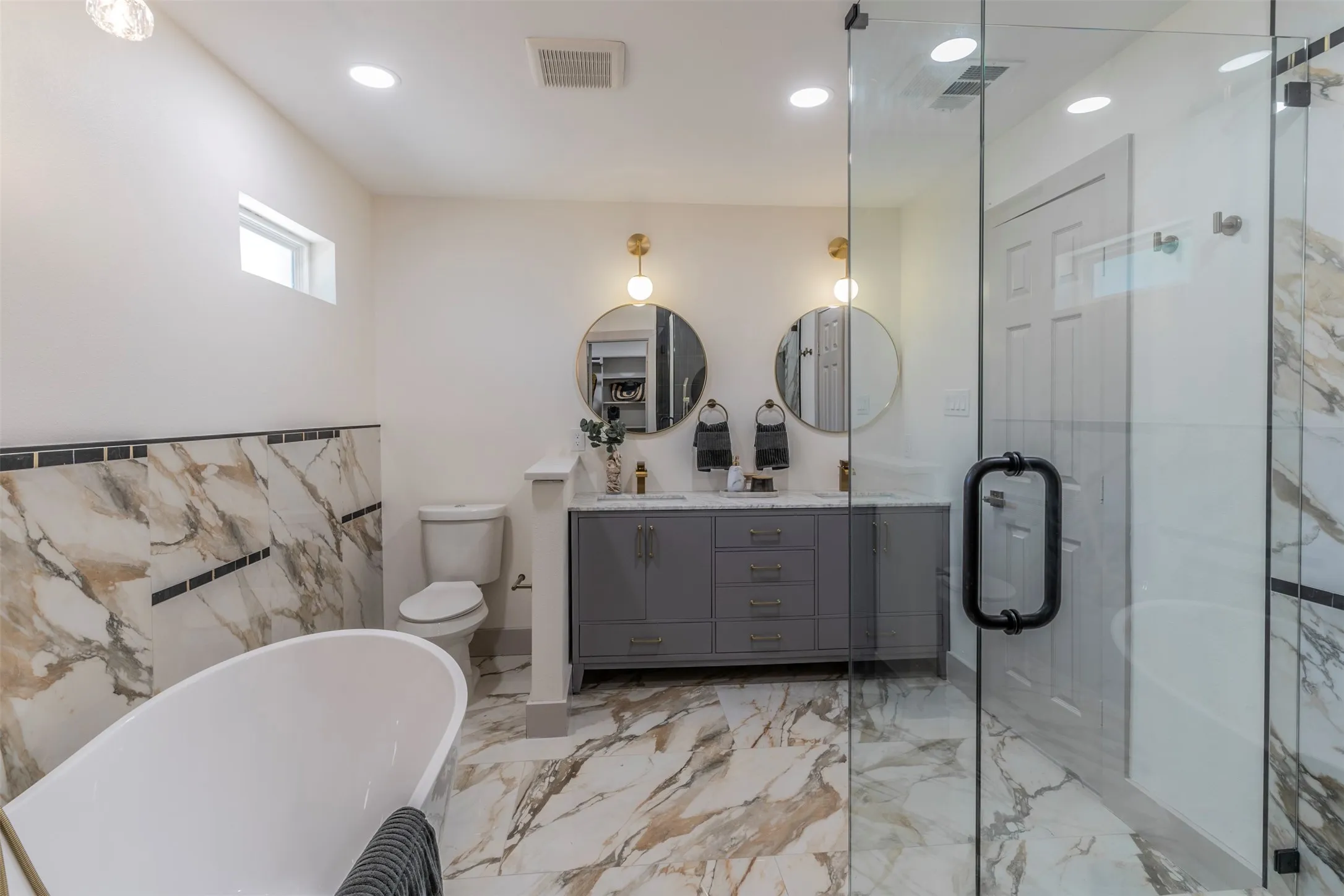Freestanding soaking tub, walk-in glass shower, dual vanity with undermount sinks, round mirrors with sconce lighting, and large format floor and wall tile
