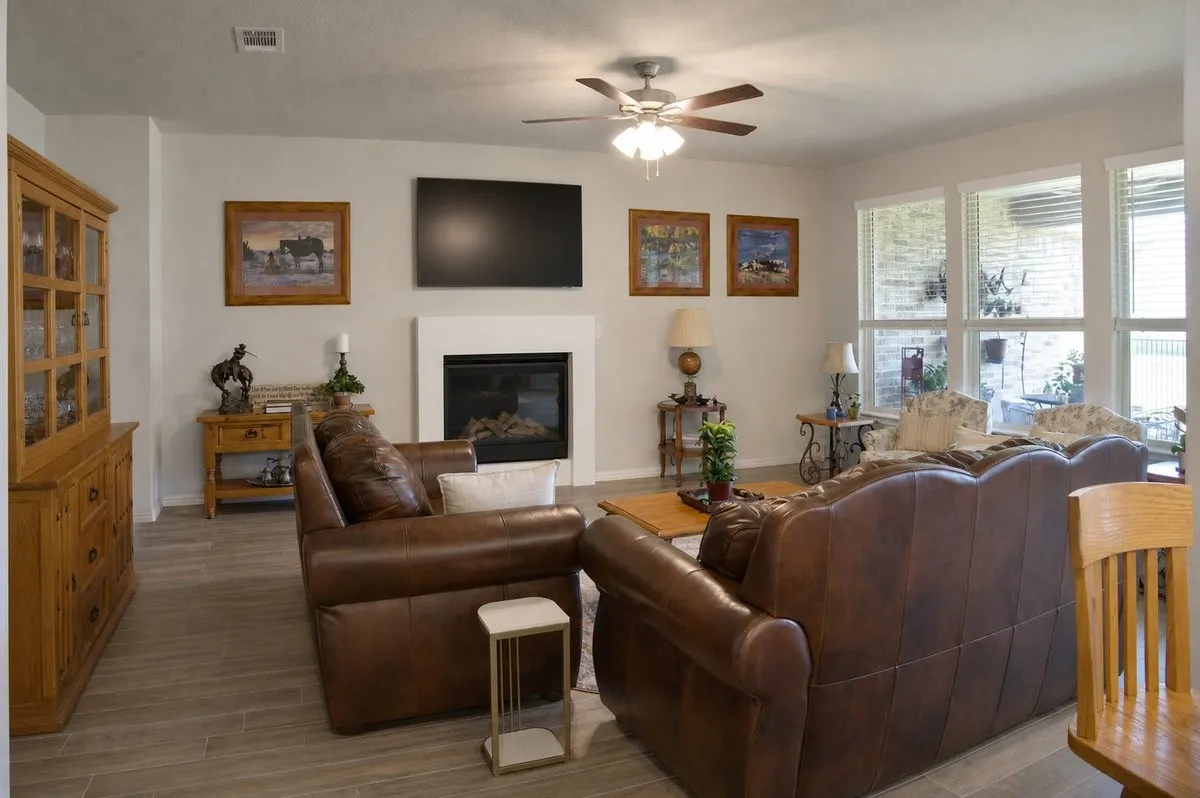 Living area featuring wood-finish flooring, a fireplace with a white mantel, a ceiling fan, and multiple windows