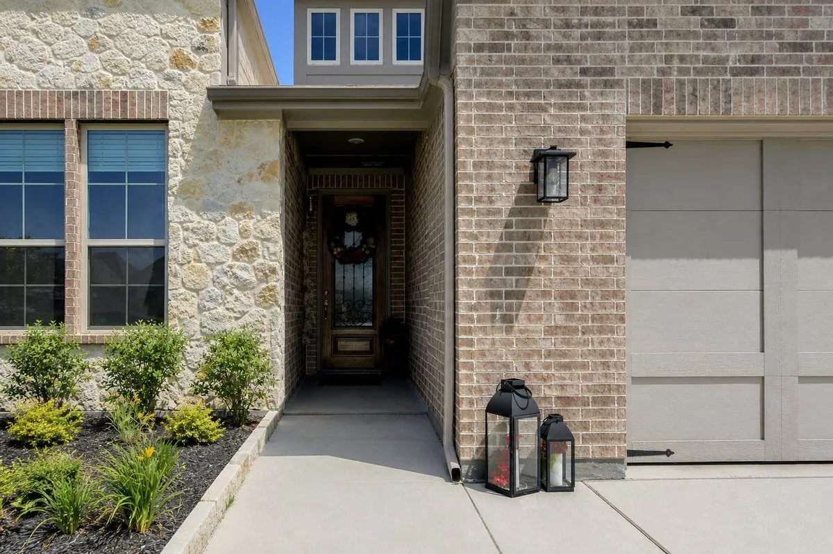 Exterior featuring a brick and stone facade, a front entry with a wood-finish door, a concrete pathway, a landscaped planting bed, and a two-car garage