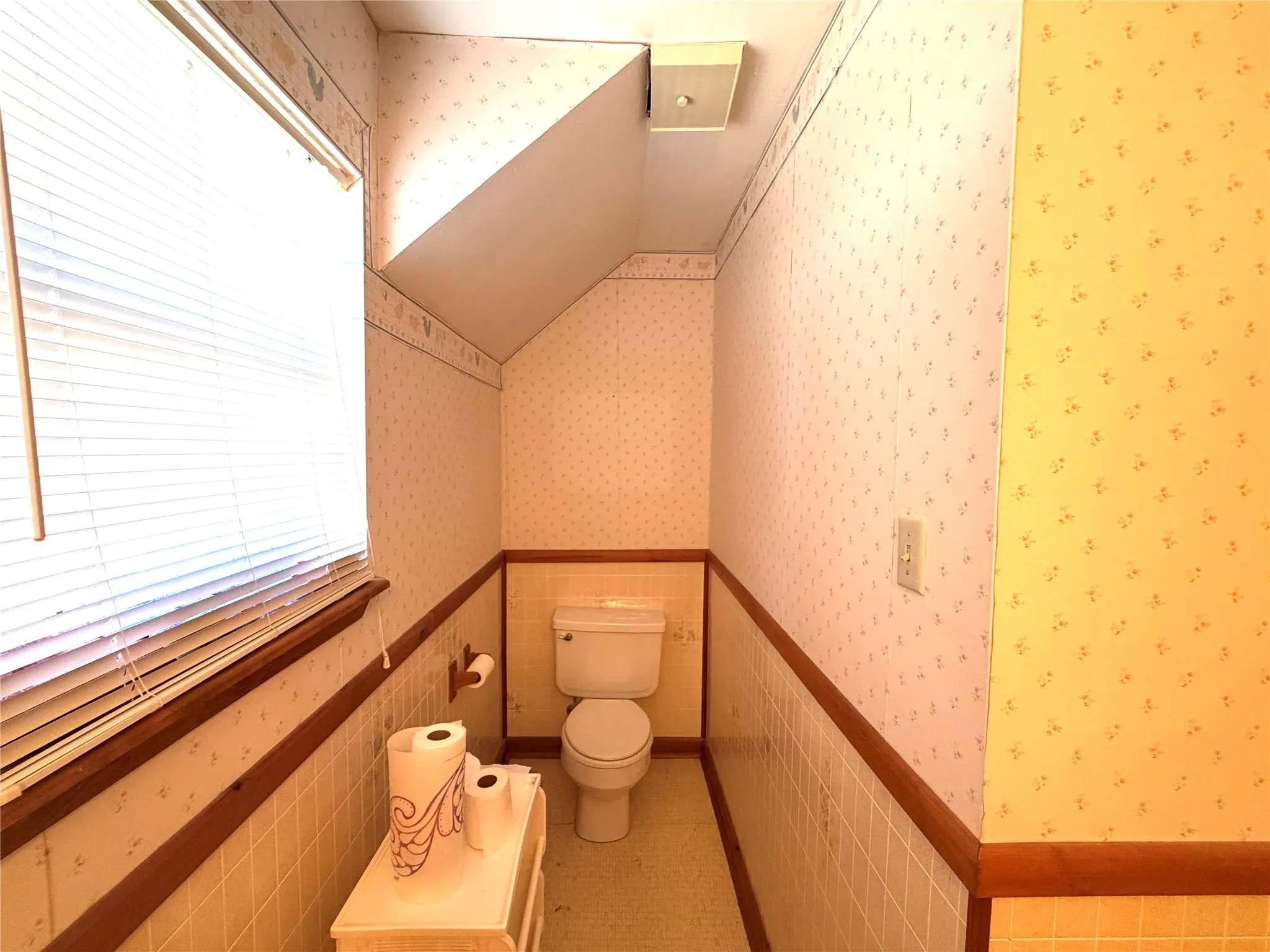 Recessed lighting fixture, window with blinds, toilet, and tiled lower walls with wood trim