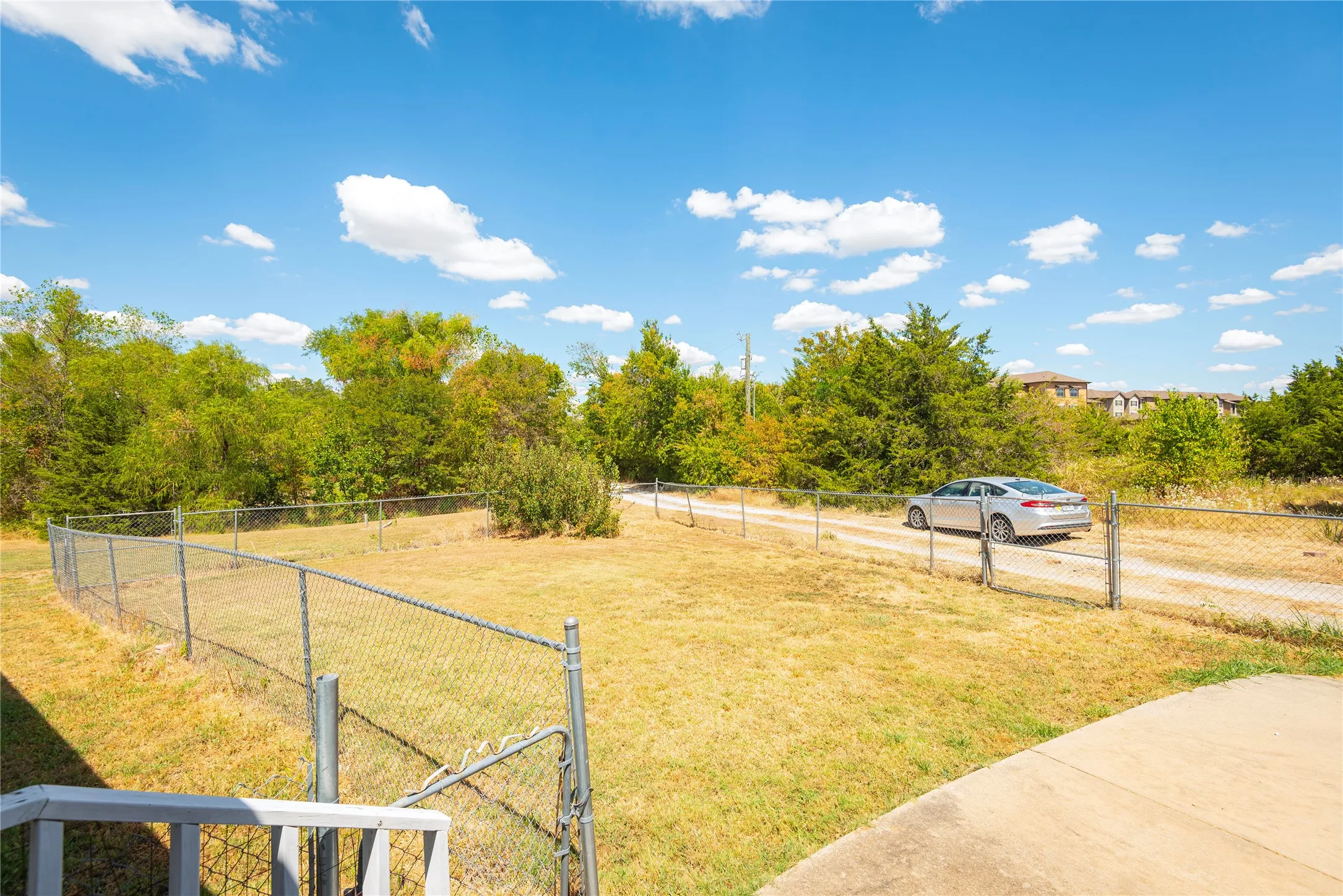 Expansive grassy area featuring chain-link fencing, a gravel driveway, and mature trees