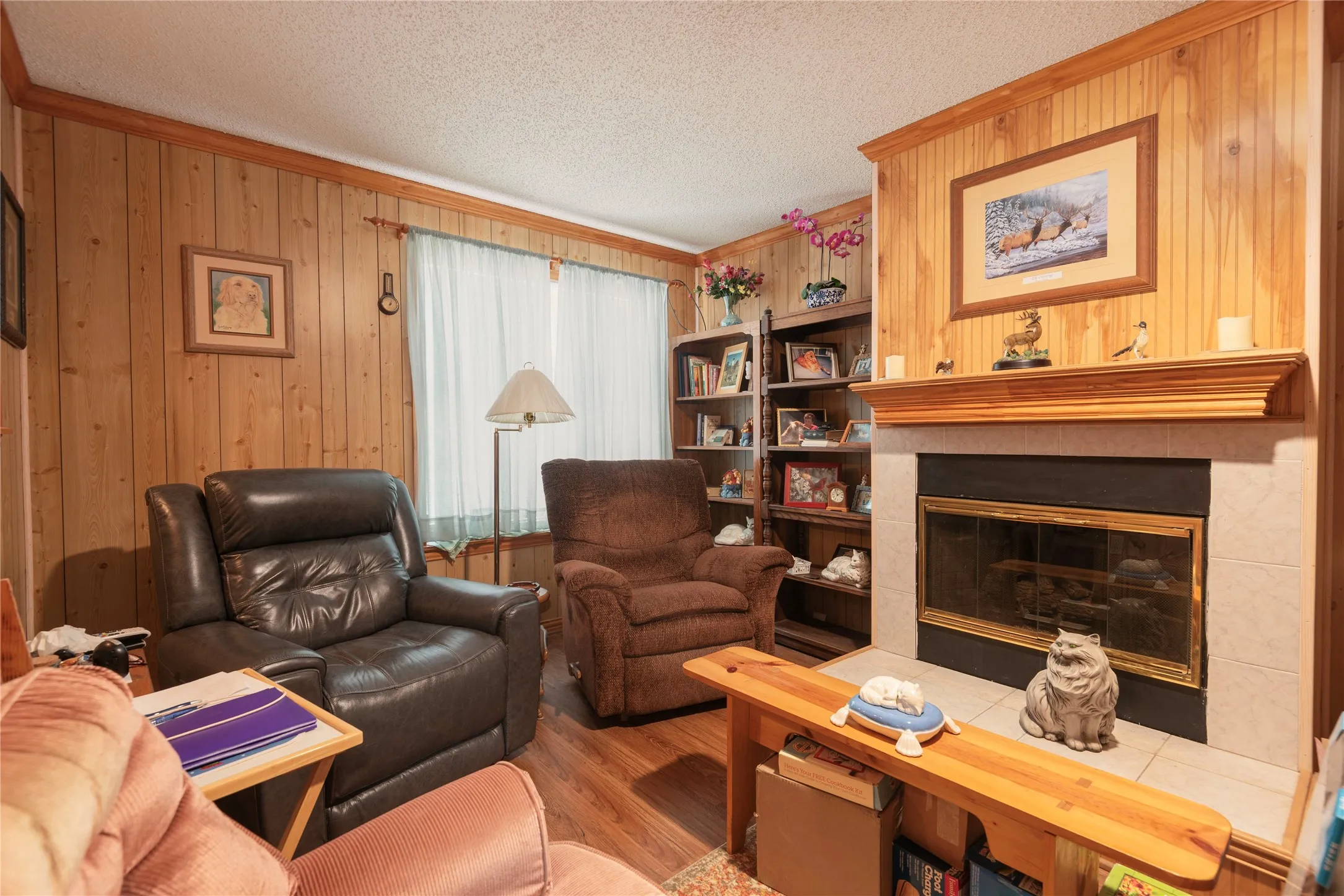 Wood-finish wall paneling, wood-finish flooring, built-in shelving, fireplace with tile surround and wood mantel, and window with privacy treatment
