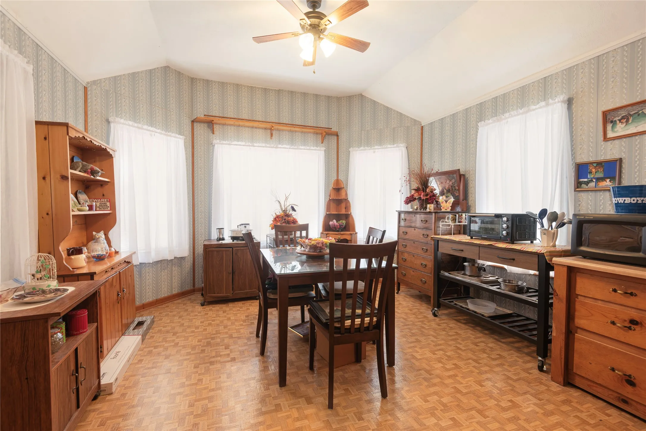 Vaulted ceiling, patterned wallpaper, wood-finish flooring, multiple windows, and a ceiling fan