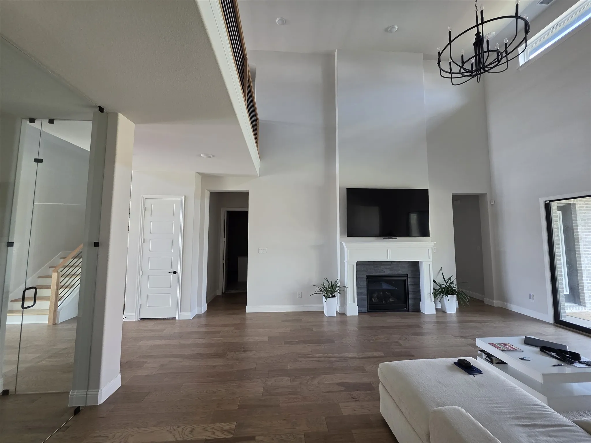 Two-story ceilings, wood-finish flooring, fireplace with white mantel and dark tile surround, and a black chandelier