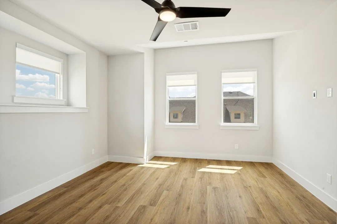 Spacious room featuring three windows, wood-finish flooring, a ceiling fan, and crisp white walls