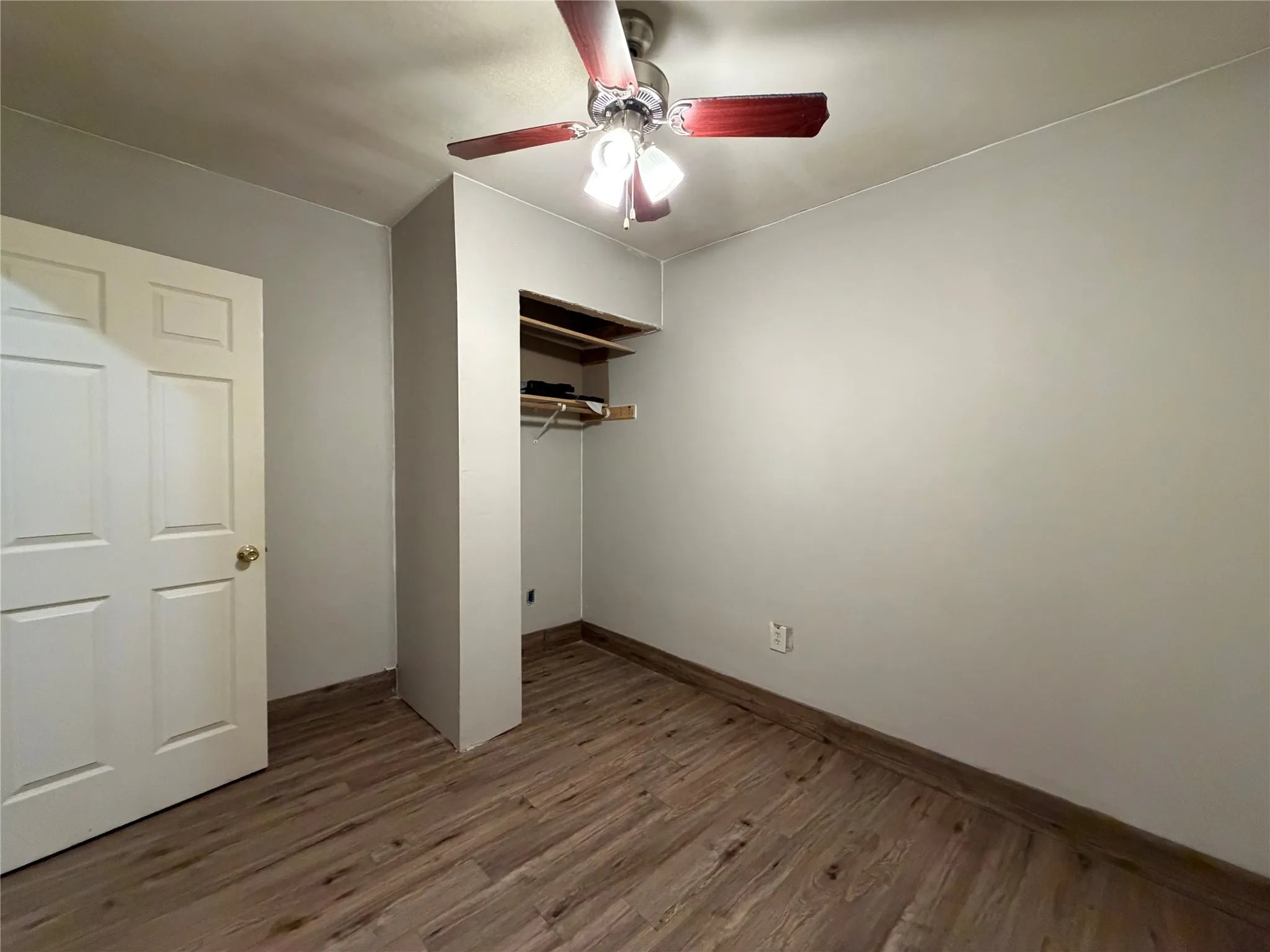 Room featuring wood-finish flooring, light gray walls, built-in open shelving, and a ceiling fan with integrated lighting