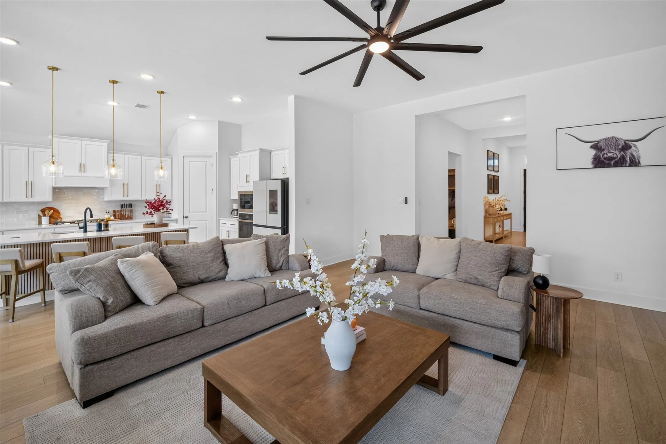 Open-concept living space featuring wood-finish flooring, a modern multi-blade ceiling fan, recessed lighting, and a kitchen with white cabinetry and pendant lighting