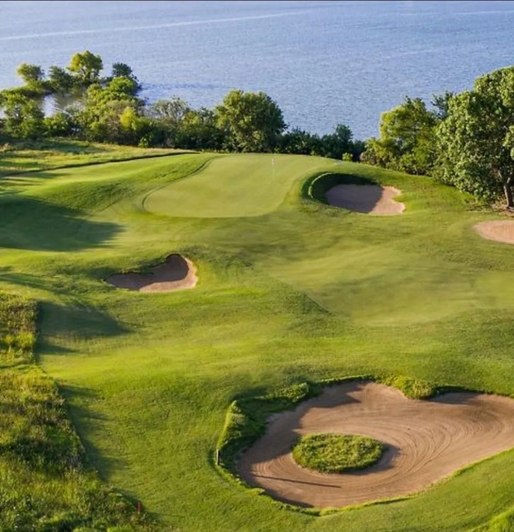 Expansive golf course featuring multiple sand traps, manicured greens, and mature trees, with a prominent water backdrop