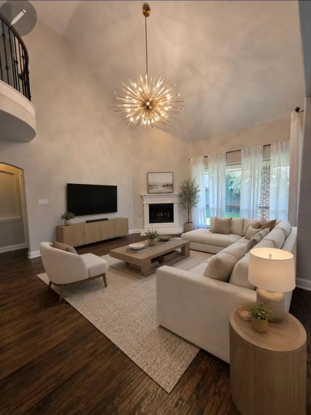 Vaulted ceiling with an ornate chandelier, fireplace with a mantle, large windows with sheer coverings, wood-finish flooring, and a curved balcony with dark metal railing