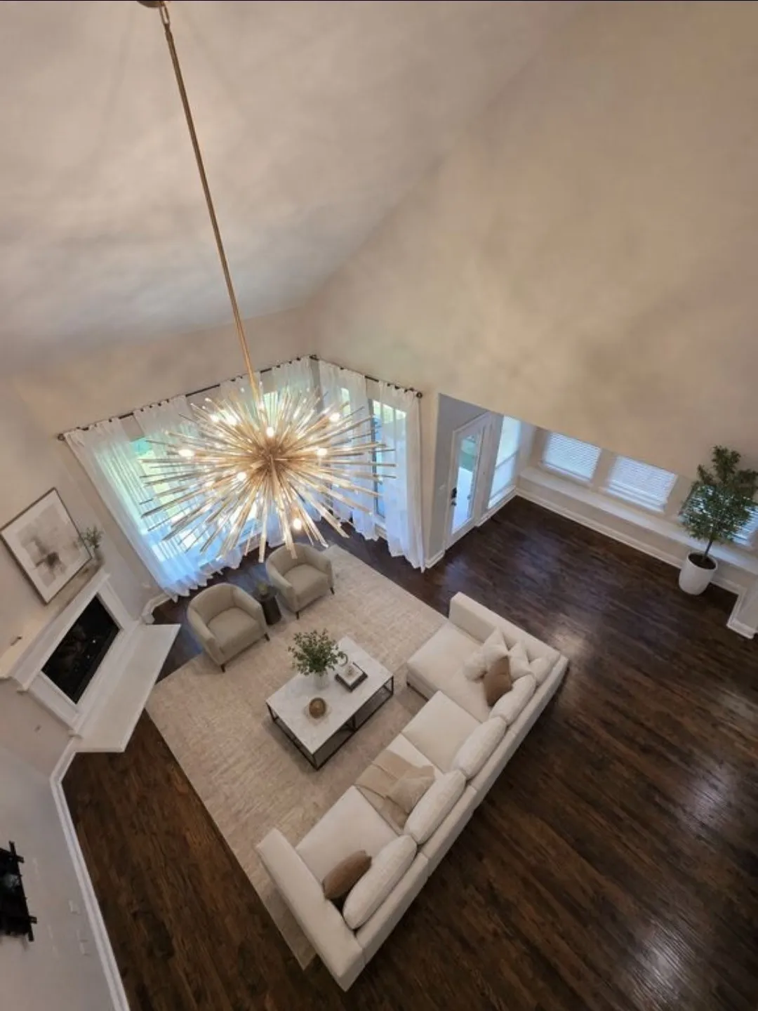 Vaulted ceiling with an elaborate chandelier, dark wood-finish flooring, white fireplace with mantel, and large windows with transom details