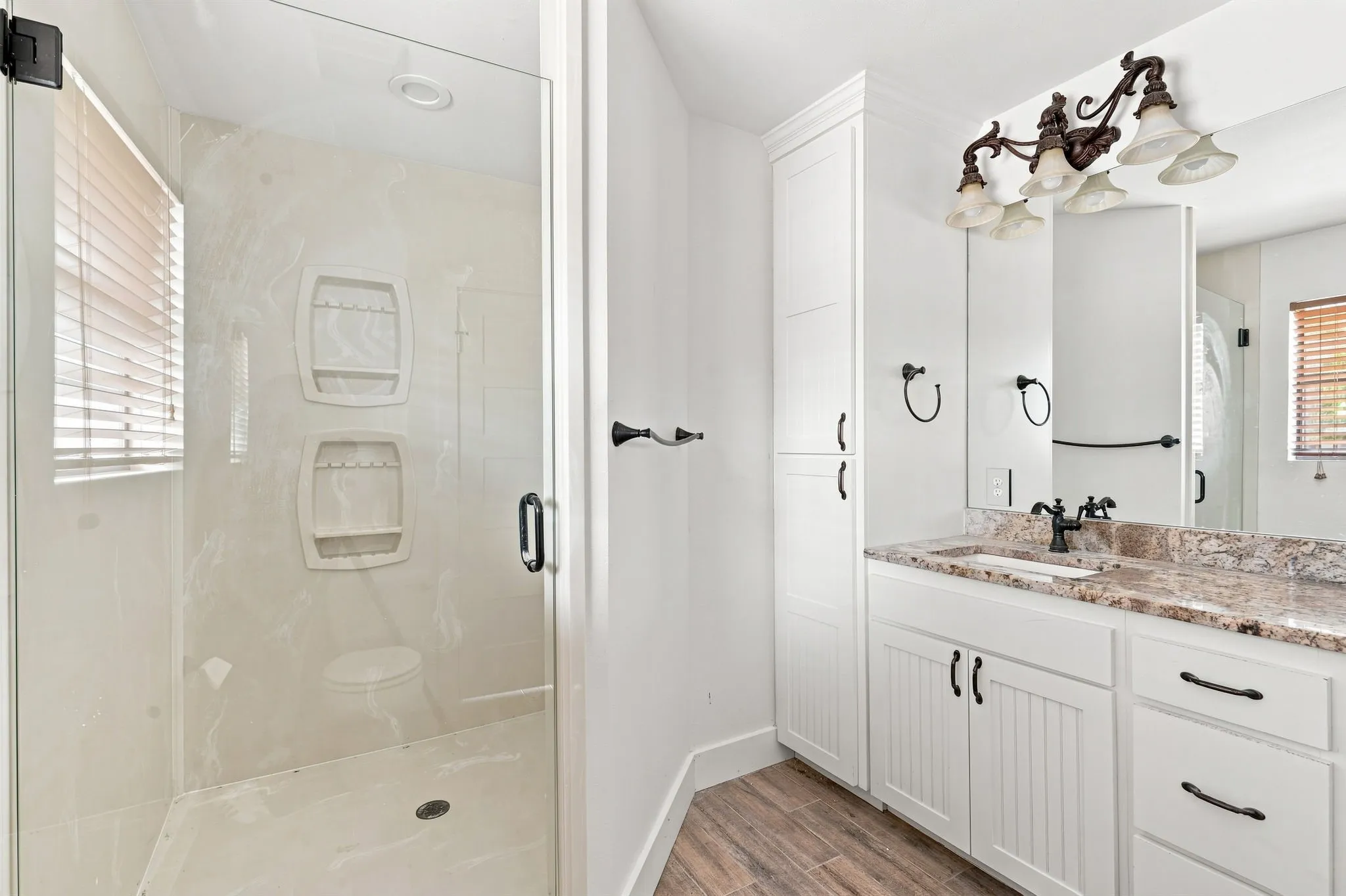 Glass-enclosed shower with tile surround, wood-finish flooring, white shaker-style cabinetry, and a vanity with a stone countertop