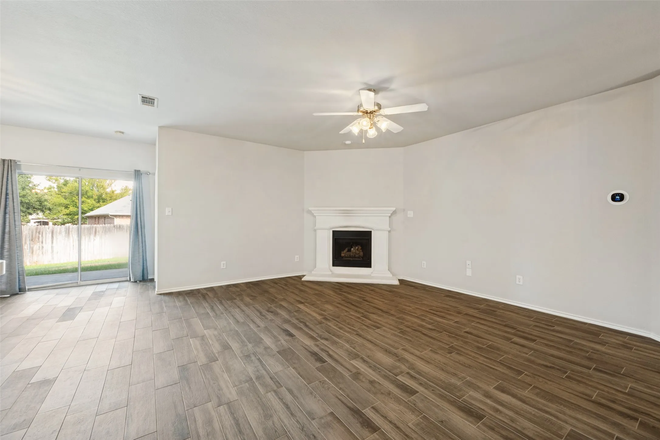 Wood-finish flooring, corner fireplace with white mantel, ceiling fan, and sliding glass door