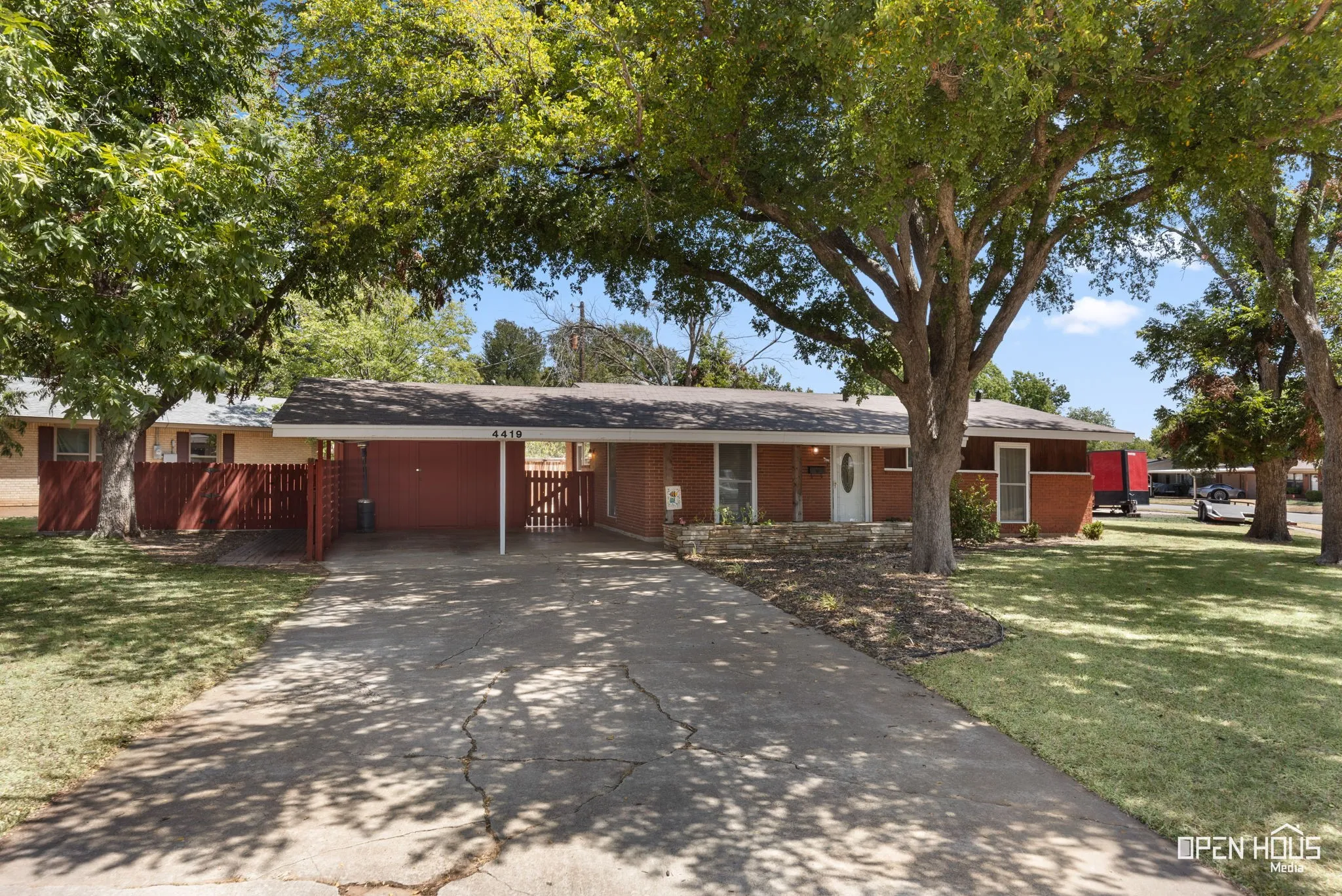 Brick exterior, attached carport, white entry door, front yard with mature trees, and a decorative rock bed