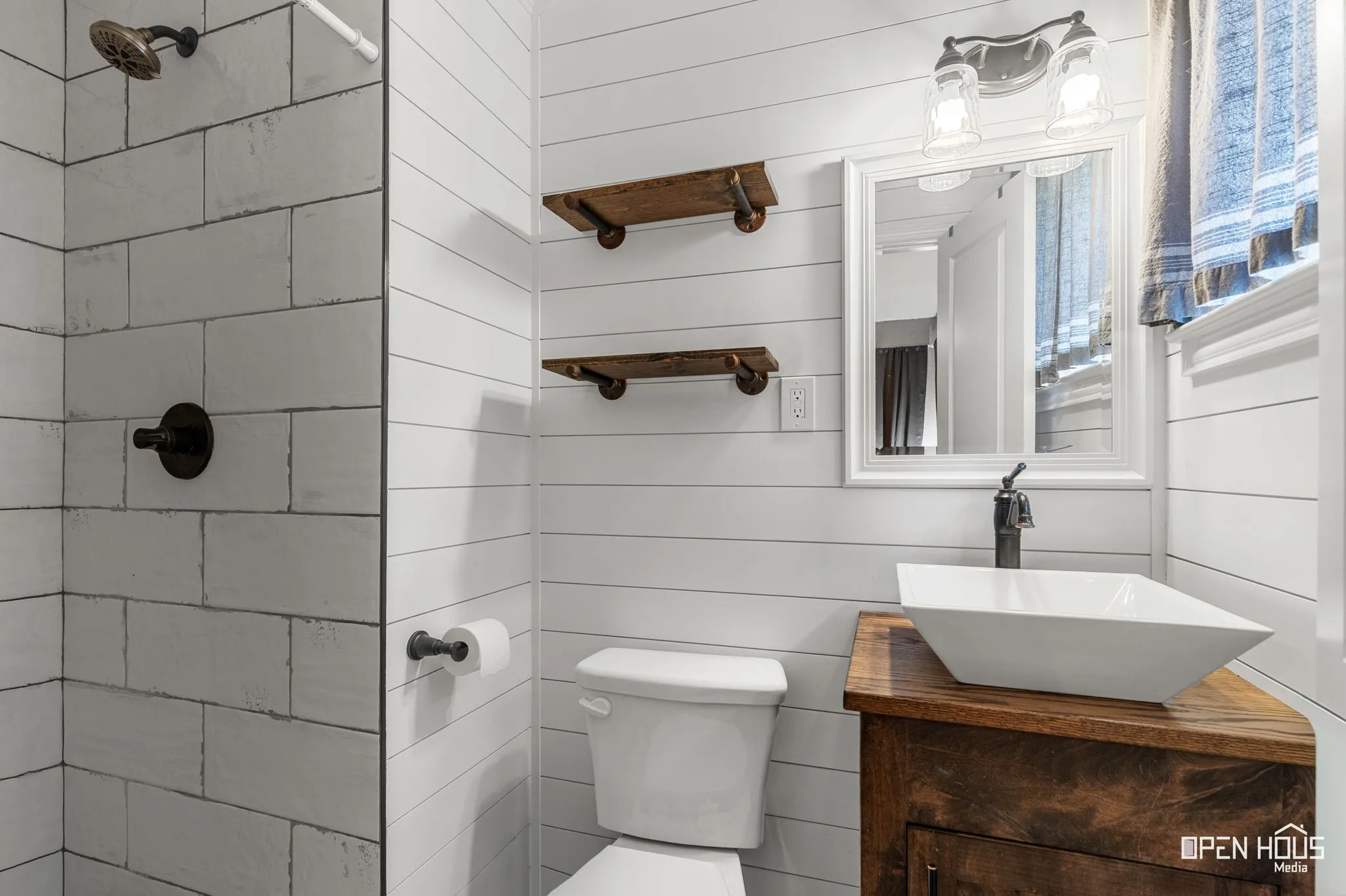 Shower with subway tile surround and dark bronze fixtures, shiplap wall paneling, wood-finish vanity with vessel sink, and wall-mounted mirror with sconce lighting