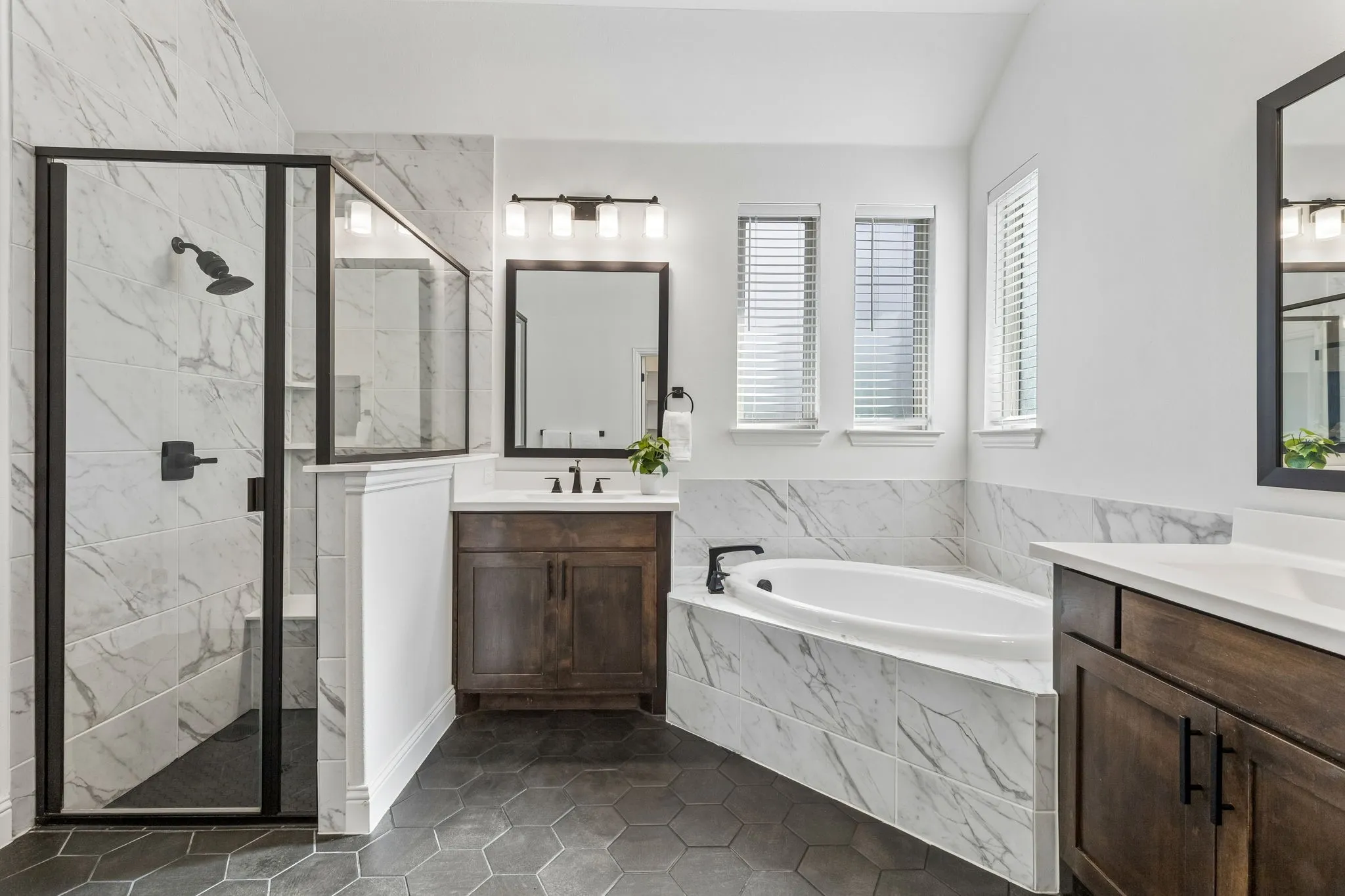 Hexagonal tile flooring, walk-in shower with black-framed glass enclosure, built-in soaking tub, dual wood-finish vanity with countertop, and double-paned windows