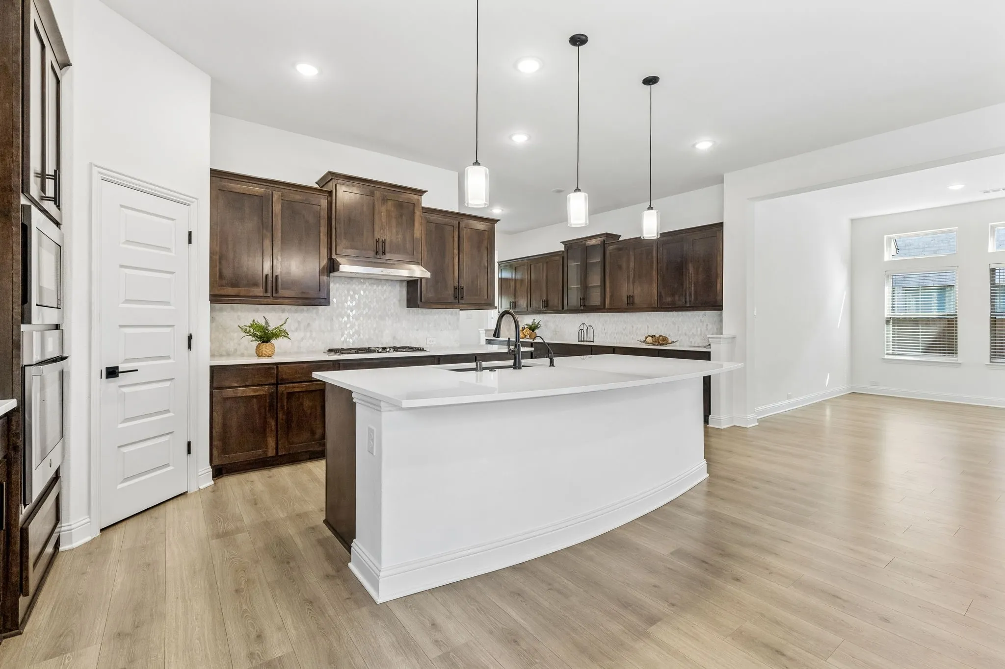 Kitchen featuring dark wood cabinetry, white countertops, tile backsplash, built-in ovens, and a center island with sink