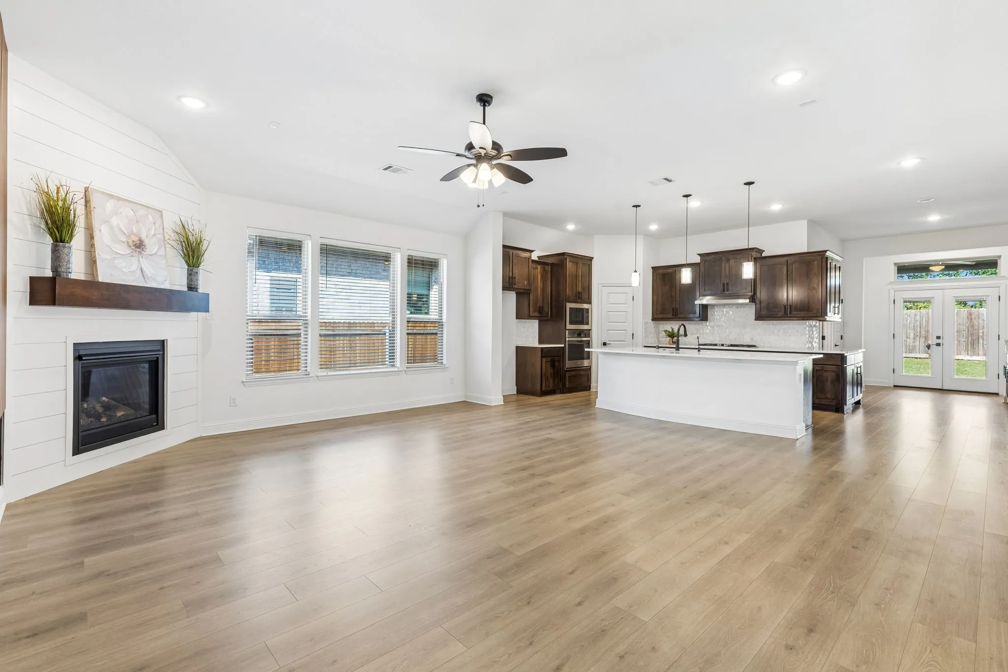 Spacious room featuring wood-finish flooring, a fireplace with a wood mantel, a kitchen island with pendant lighting, dark wood cabinetry, and double glass-paneled doors