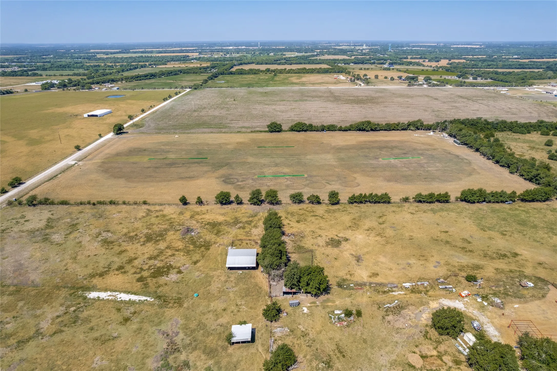 Expansive agricultural land featuring a gravel road, multiple outbuildings, and mature trees defining property lines