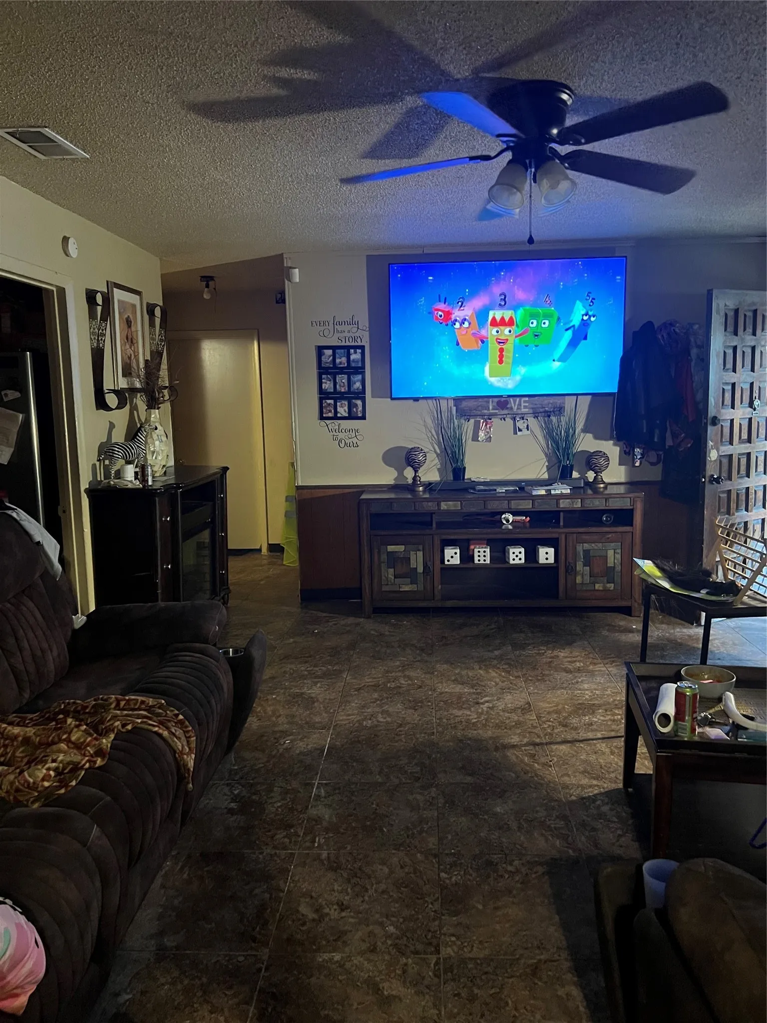 Textured tile flooring throughout, featuring a ceiling fan with integrated lighting, and a wall-mounted entertainment console