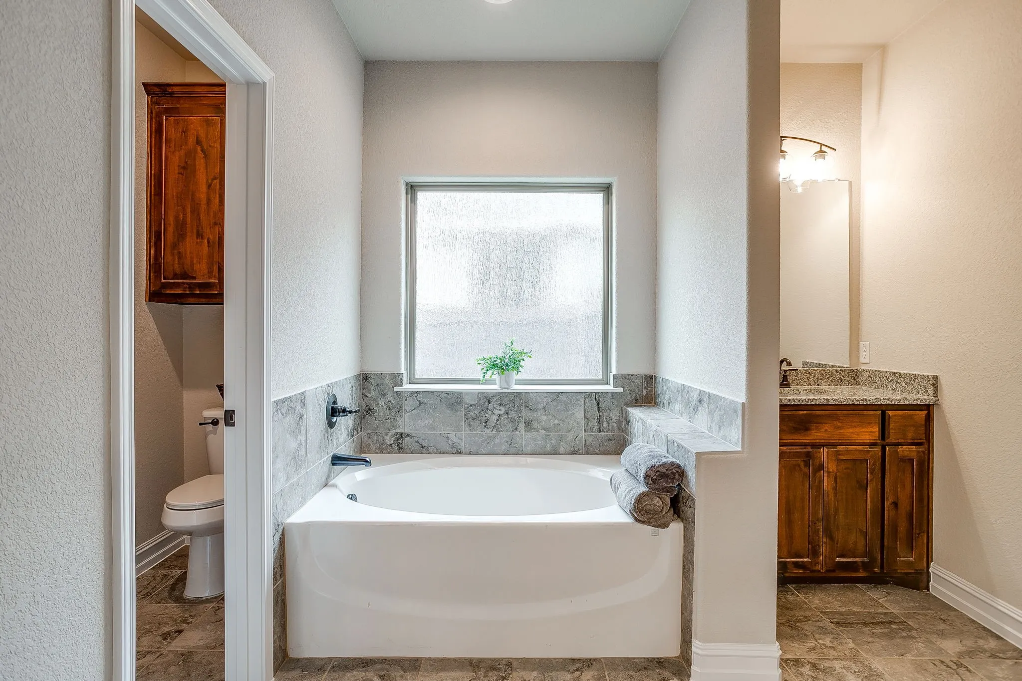 Built-in soaking tub with tile surround, framed window, separate water closet, and vanity with granite countertop