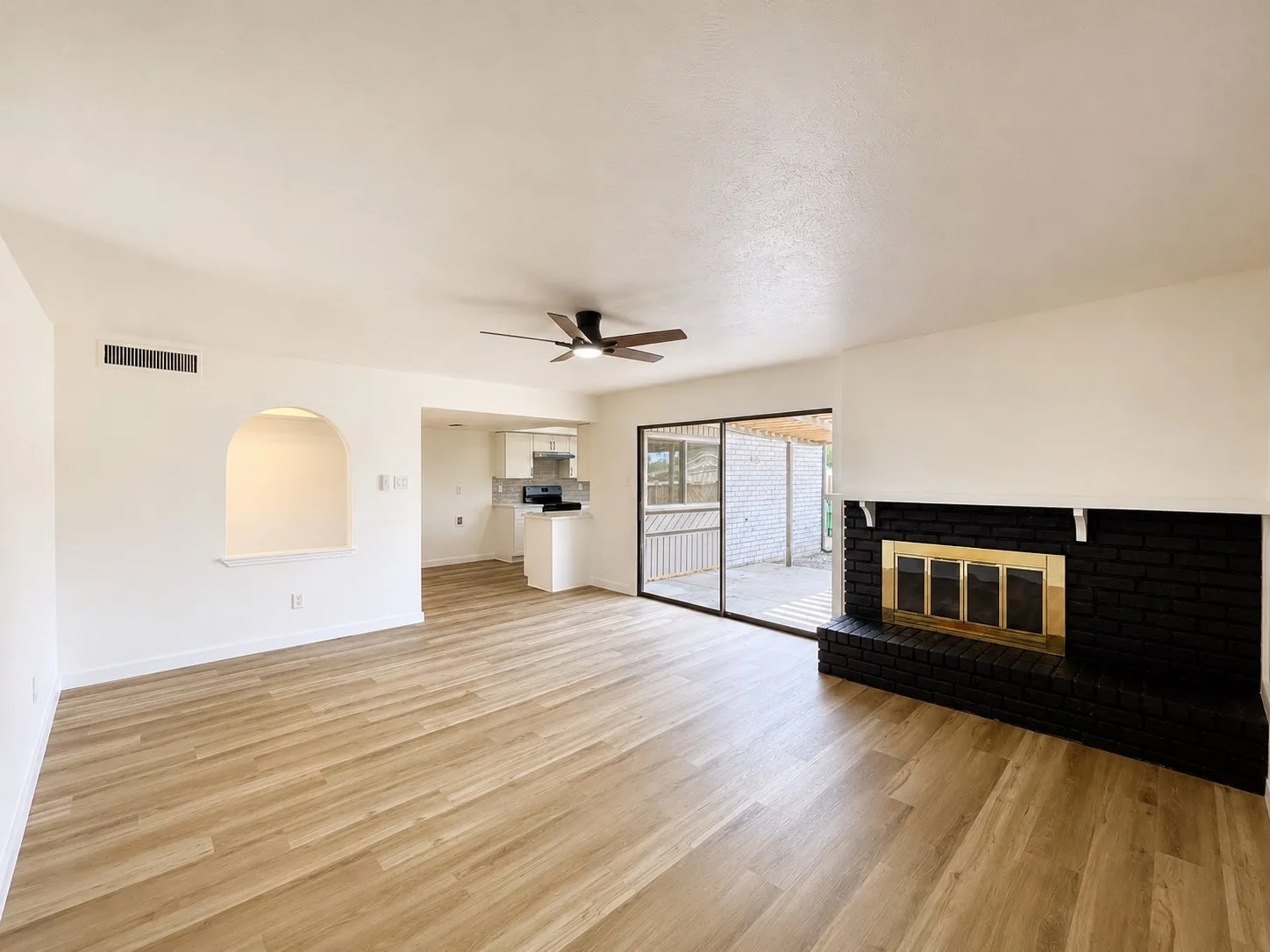 Wood-finish flooring, black brick fireplace with a gold-tone insert, arched wall niche, ceiling fan, and sliding glass door