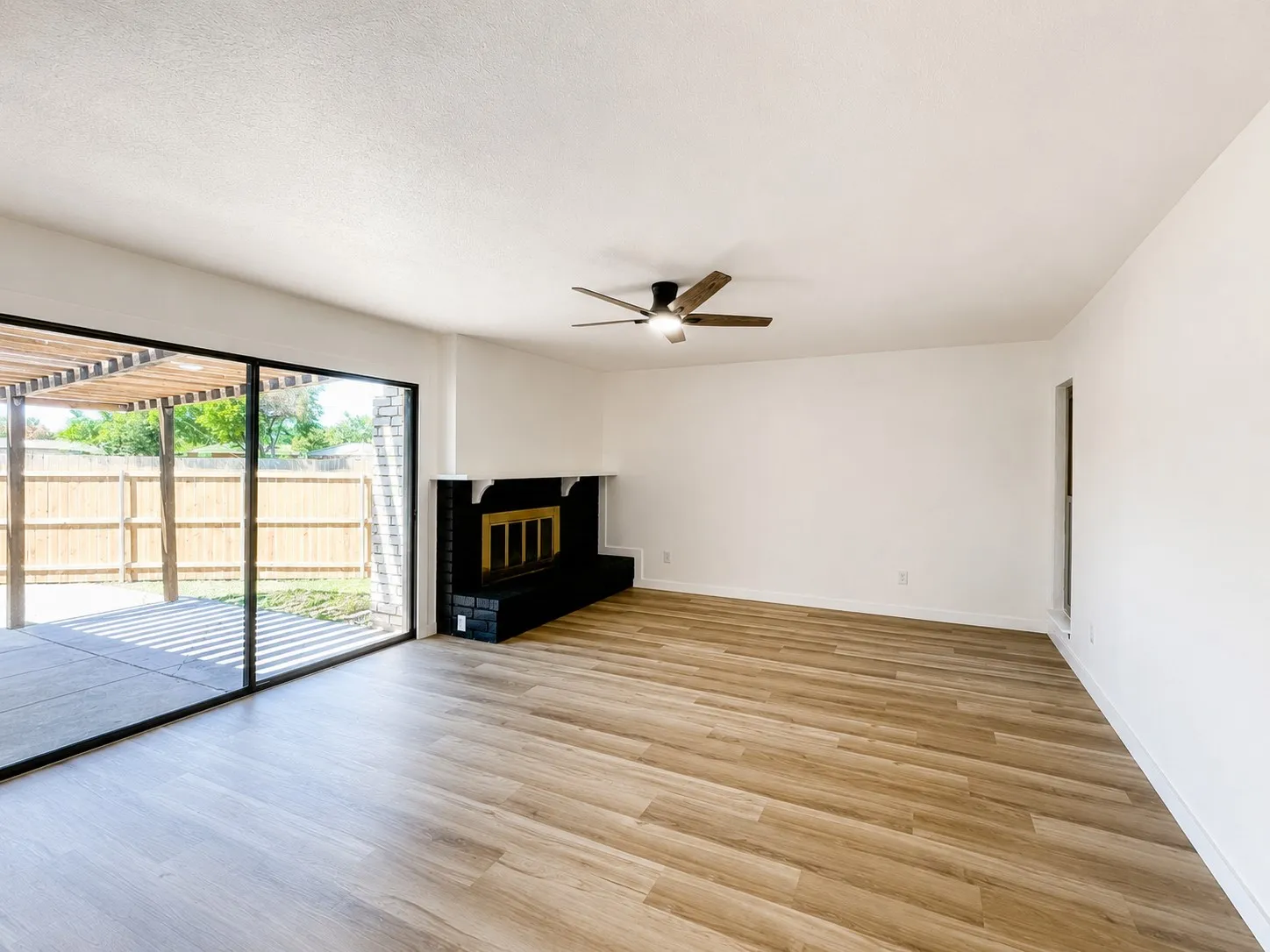 Wood-finish flooring, black brick fireplace with a mantel, ceiling fan, and large sliding glass doors leading to an outdoor patio with a pergola