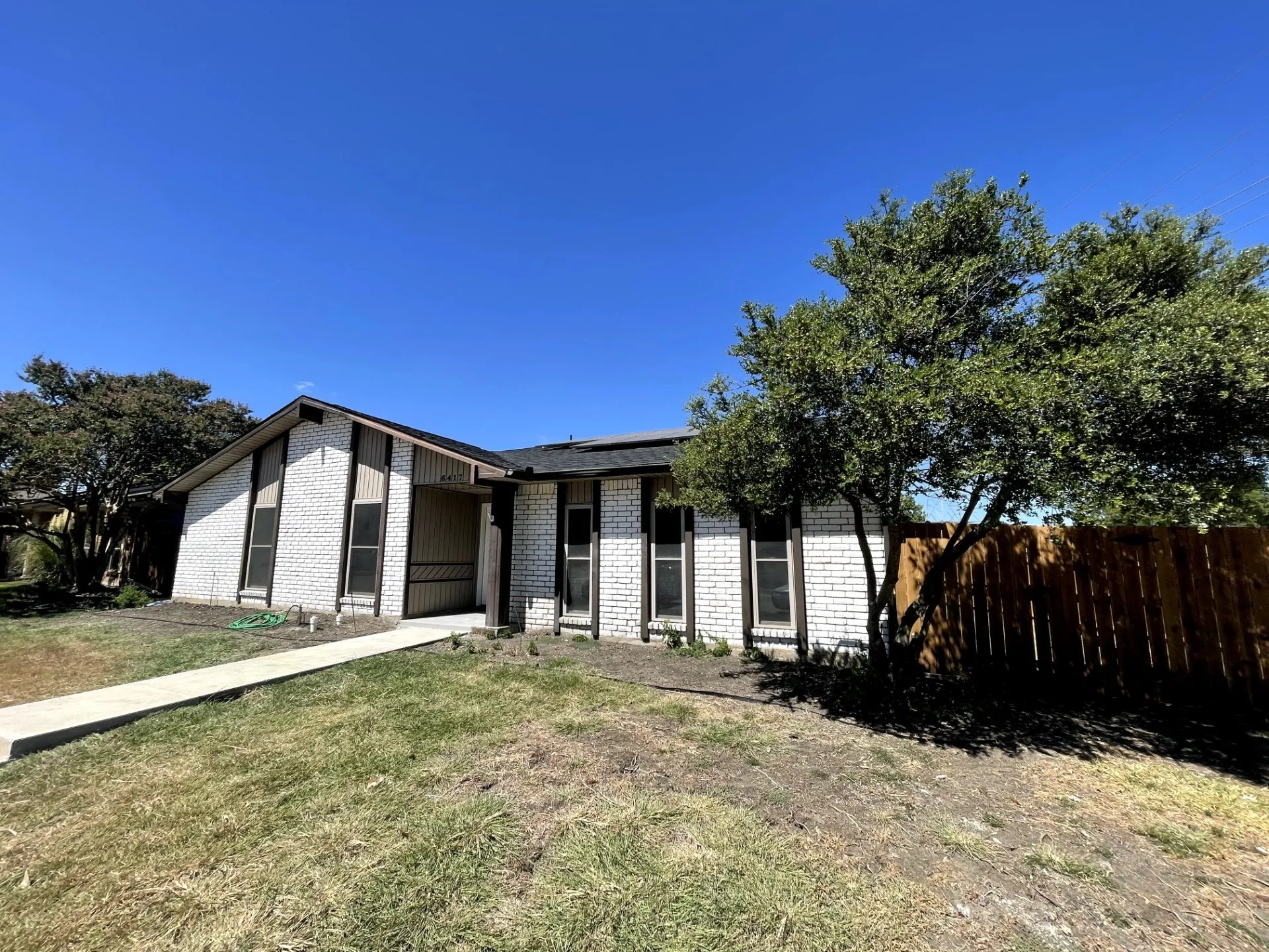 White brick exterior, dark-trimmed vertical windows, attached carport, mature front yard trees, and a concrete walkway