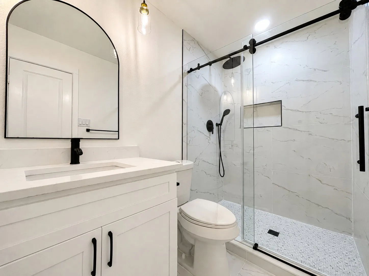 White vanity with integrated sink and matte black faucet, framed arched mirror, frameless glass shower enclosure with matte black hardware, shower niche, and white tiled walls
