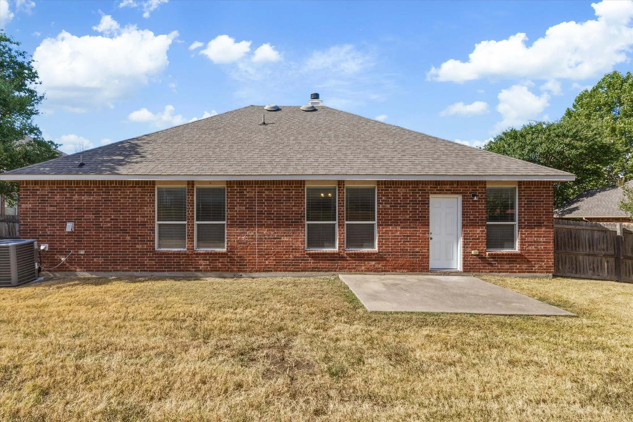 Red brick exterior with a gray shingle roof, multiple windows, and a concrete patio