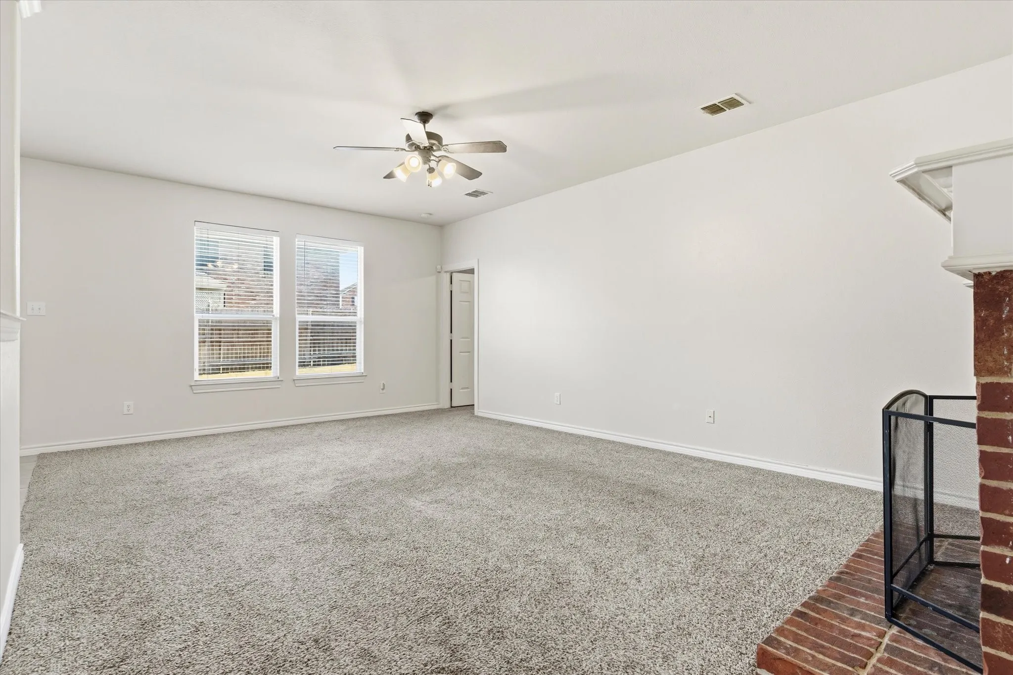 Expansive carpeting, dual windows with blinds, a ceiling fan with integrated lighting, and a brick fireplace with a decorative mantel