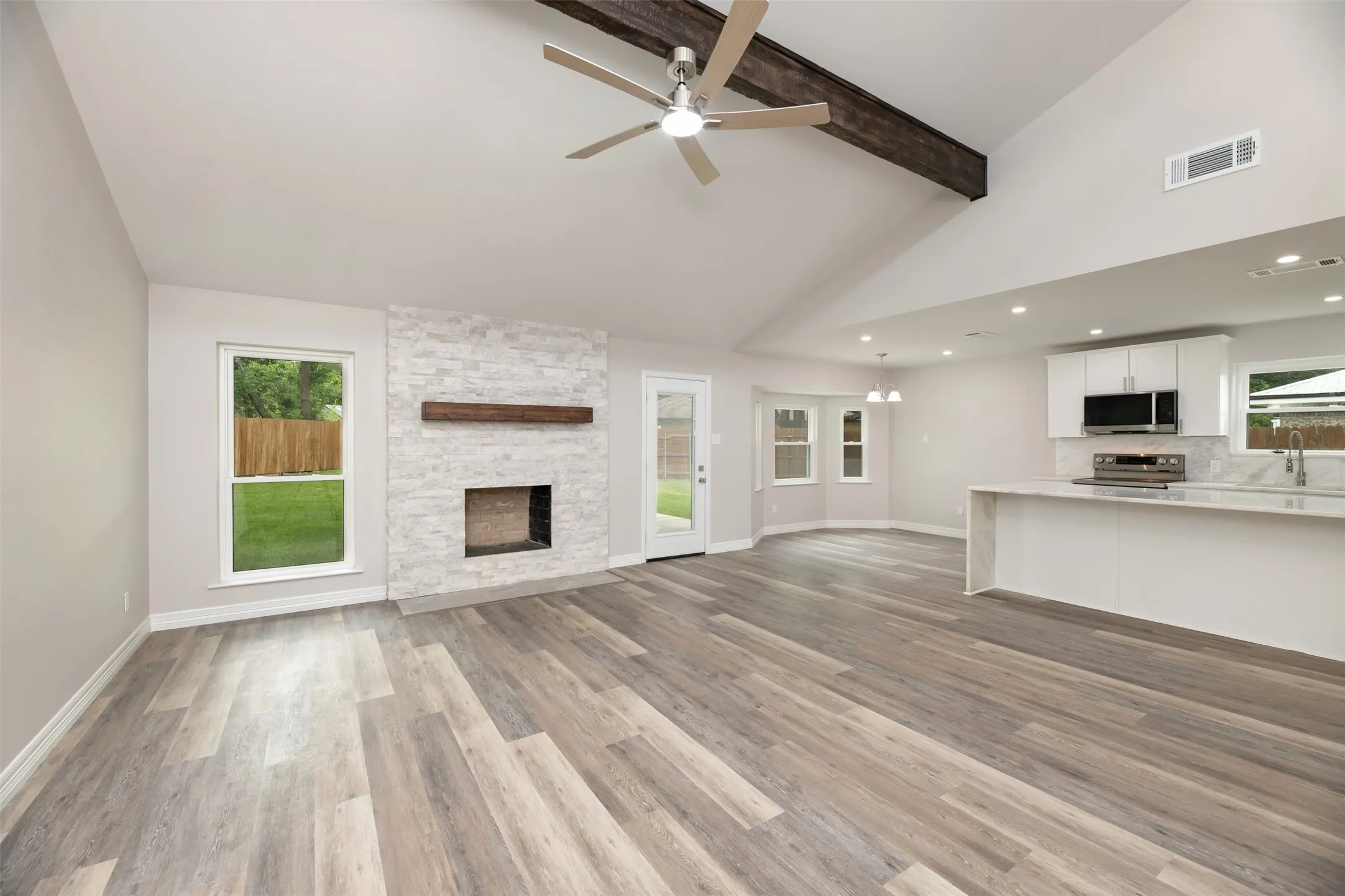 Vaulted ceiling with exposed wood beam, wood-finish flooring, stone fireplace with wood mantel, modern kitchen cabinetry, and bay windows