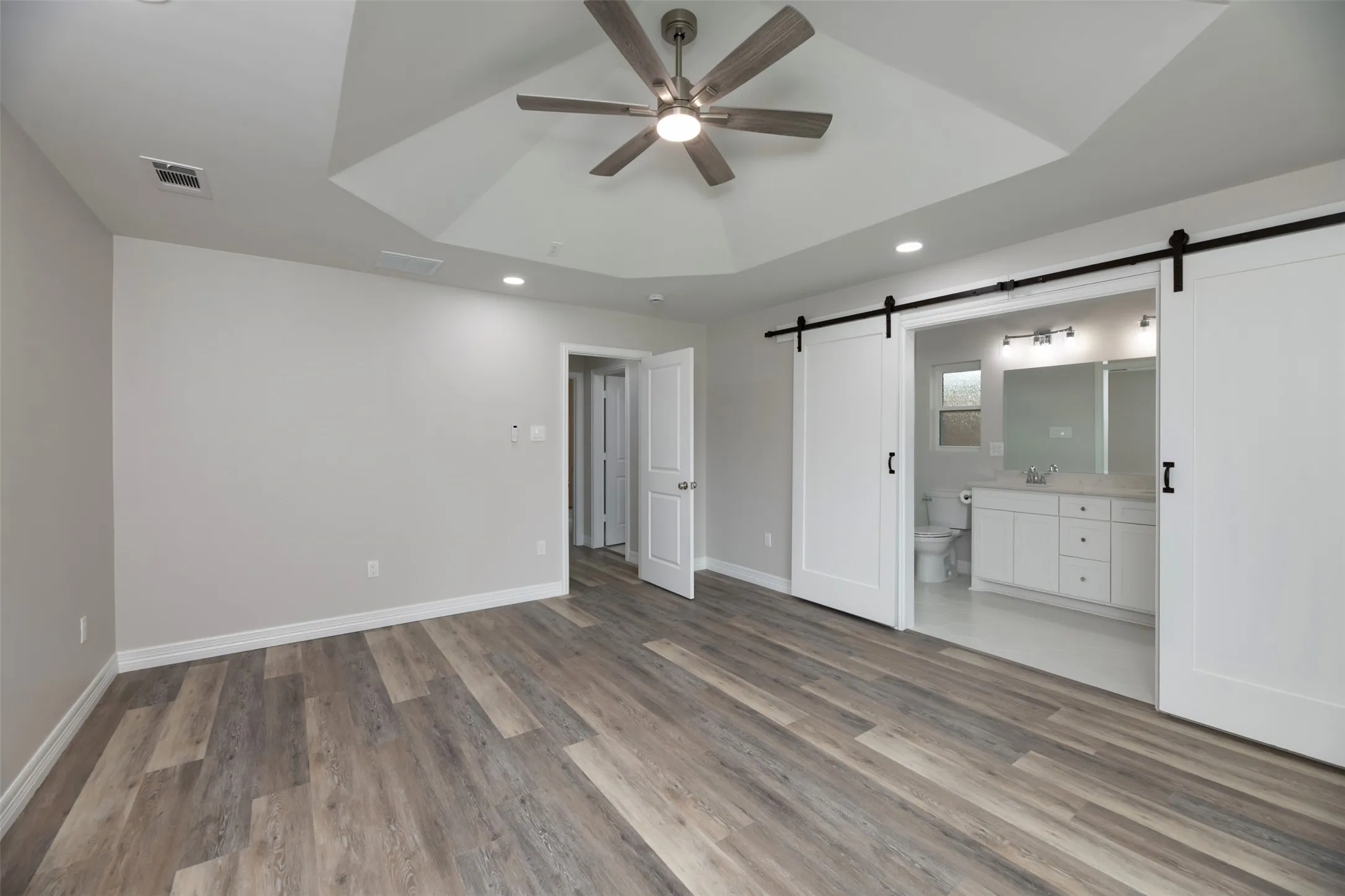 Wood-finish flooring, tray ceiling with recessed lighting, ceiling fan, and white barn doors